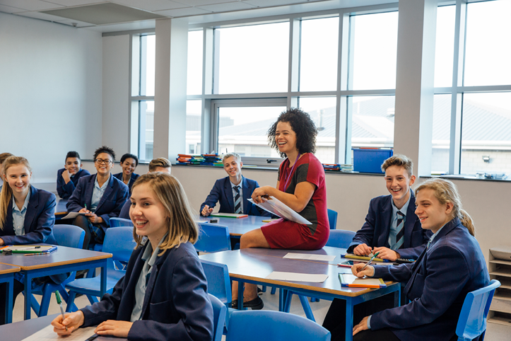 Classroom with students in blue uniforms and a teacher sitting on a desk, all smiling and engaging in a discussion.