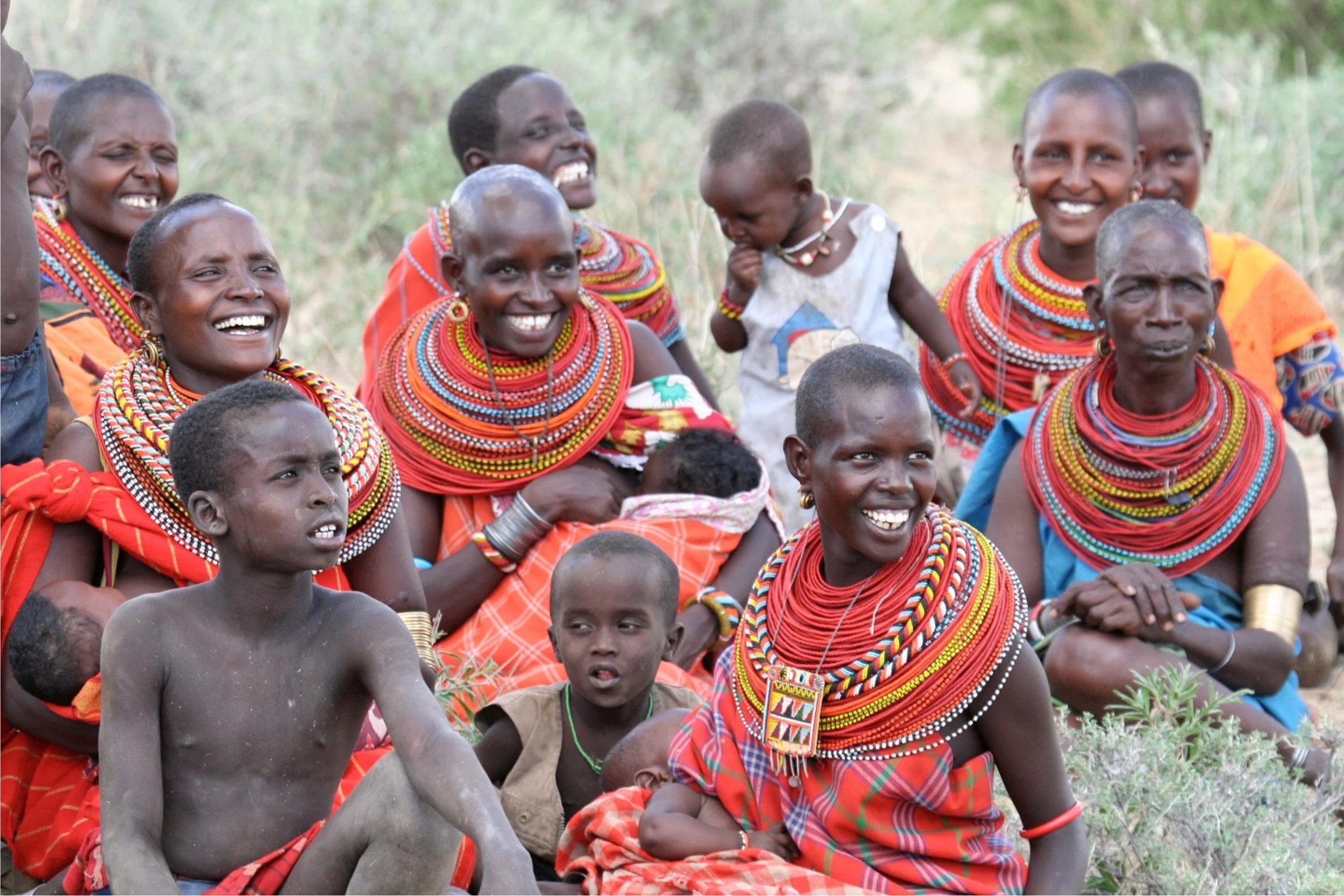 Group of Maasai women and children in traditional attire, some sitting on the ground outdoors, smiling and engaging with each other.