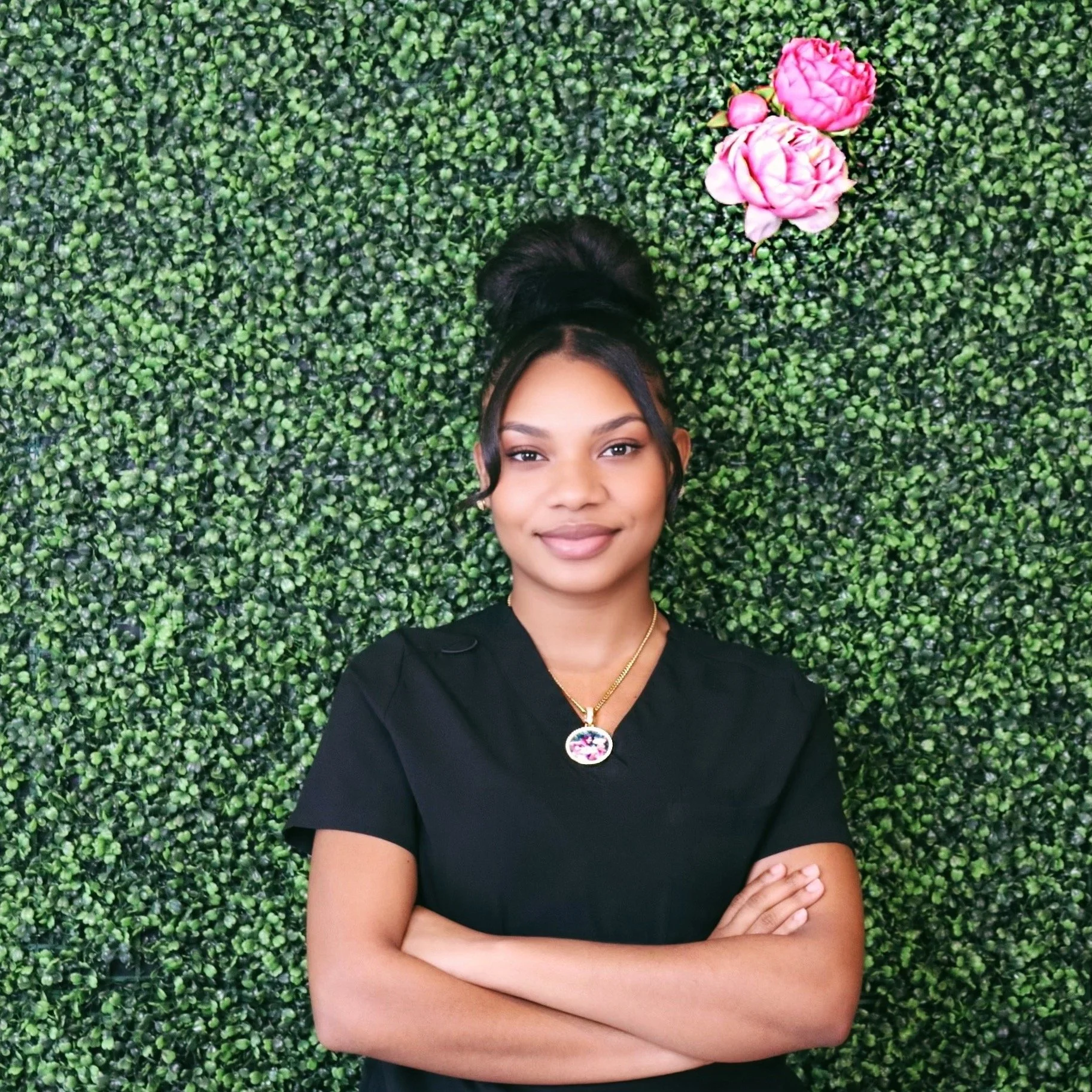 A smiling woman with arms crossed, wearing a black shirt and necklace, standing in front of a green leafy wall with pink flowers.