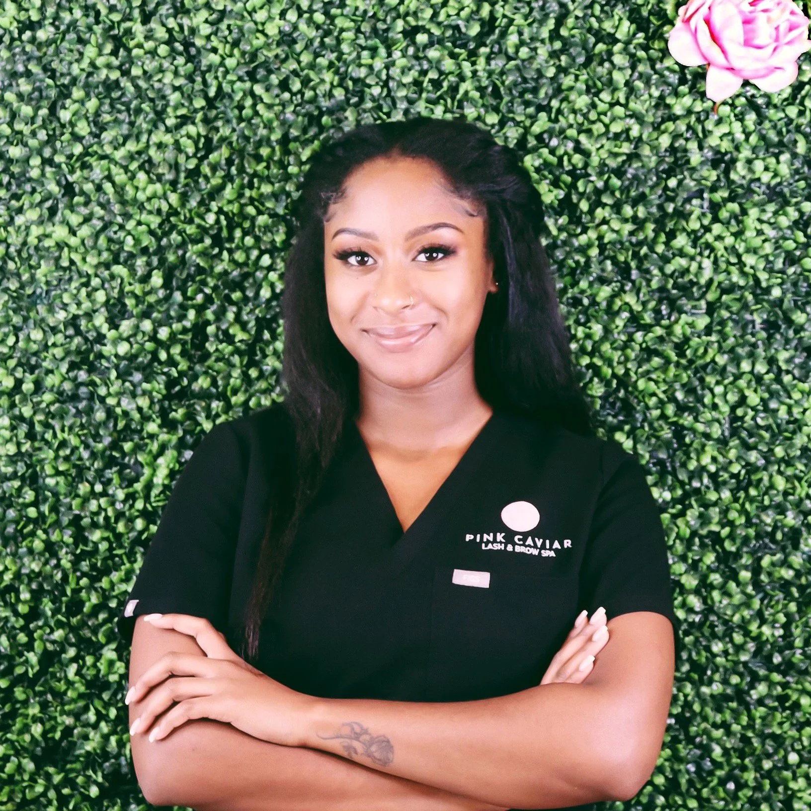A woman with long black hair standing with arms crossed in front of a green leafy wall, wearing a black uniform with a pink caviar logo on it.