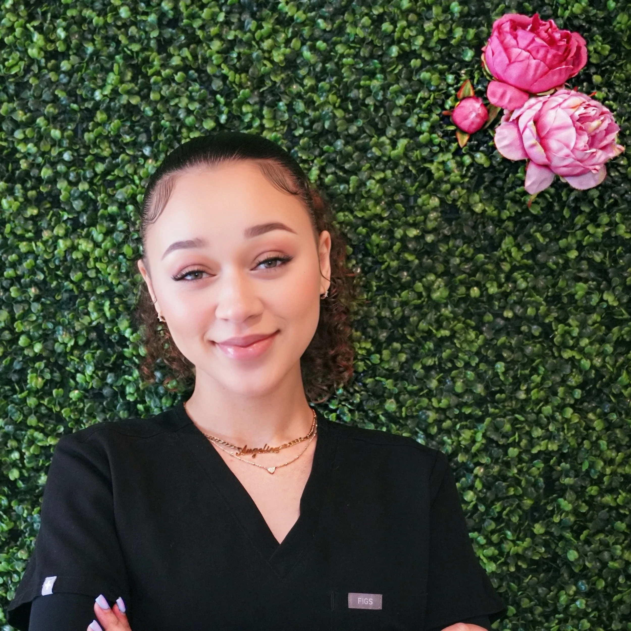 Young woman smiling with crossed arms in front of green hedge wall with pink roses, wearing black top and layered necklaces.