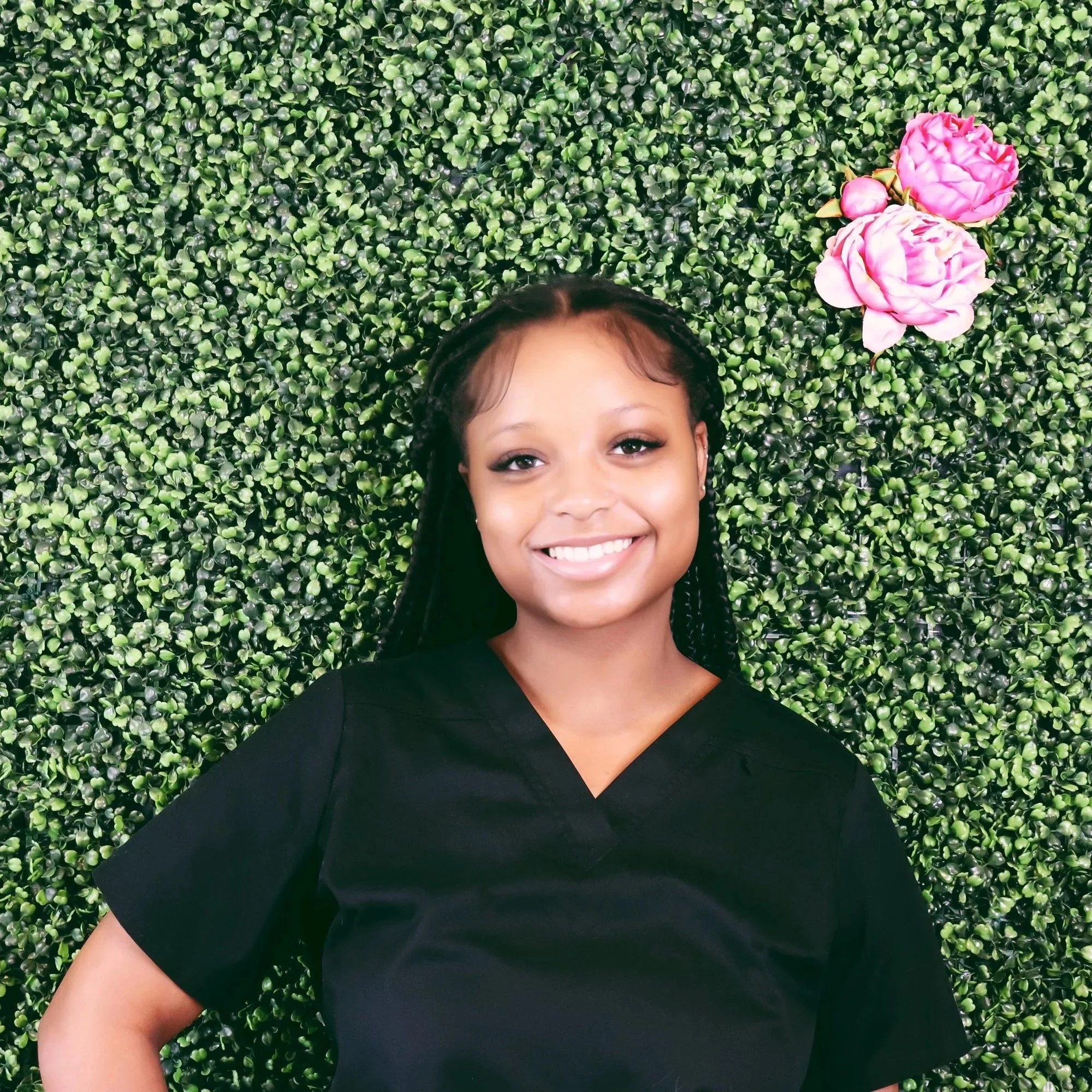 A woman in a black scrubs top smiling while lying on a green leafy backdrop with pink flowers.