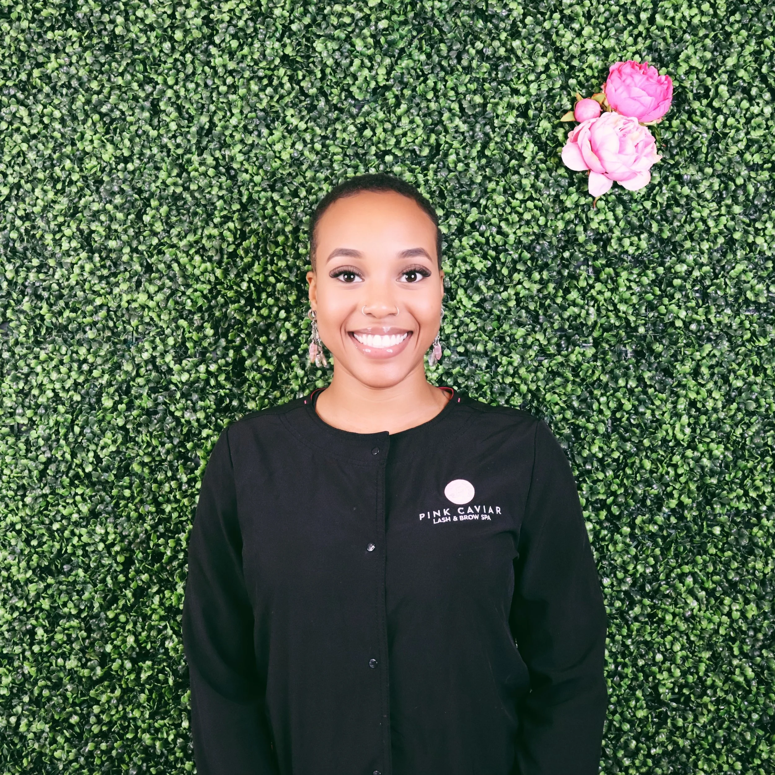 A smiling woman with earrings and a nose ring, standing in front of a lush green wall with pink flowers, wearing a black coat with a logo reading 'Pink Caviar Lash & Brow Spa'.