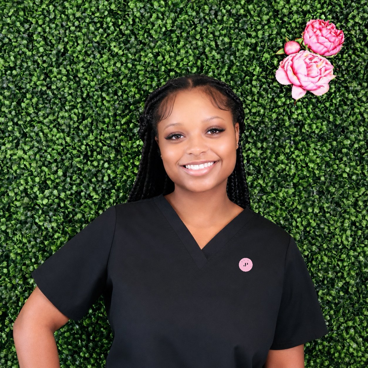 A woman in a black scrubs top smiling while lying on a green leafy backdrop with pink flowers.