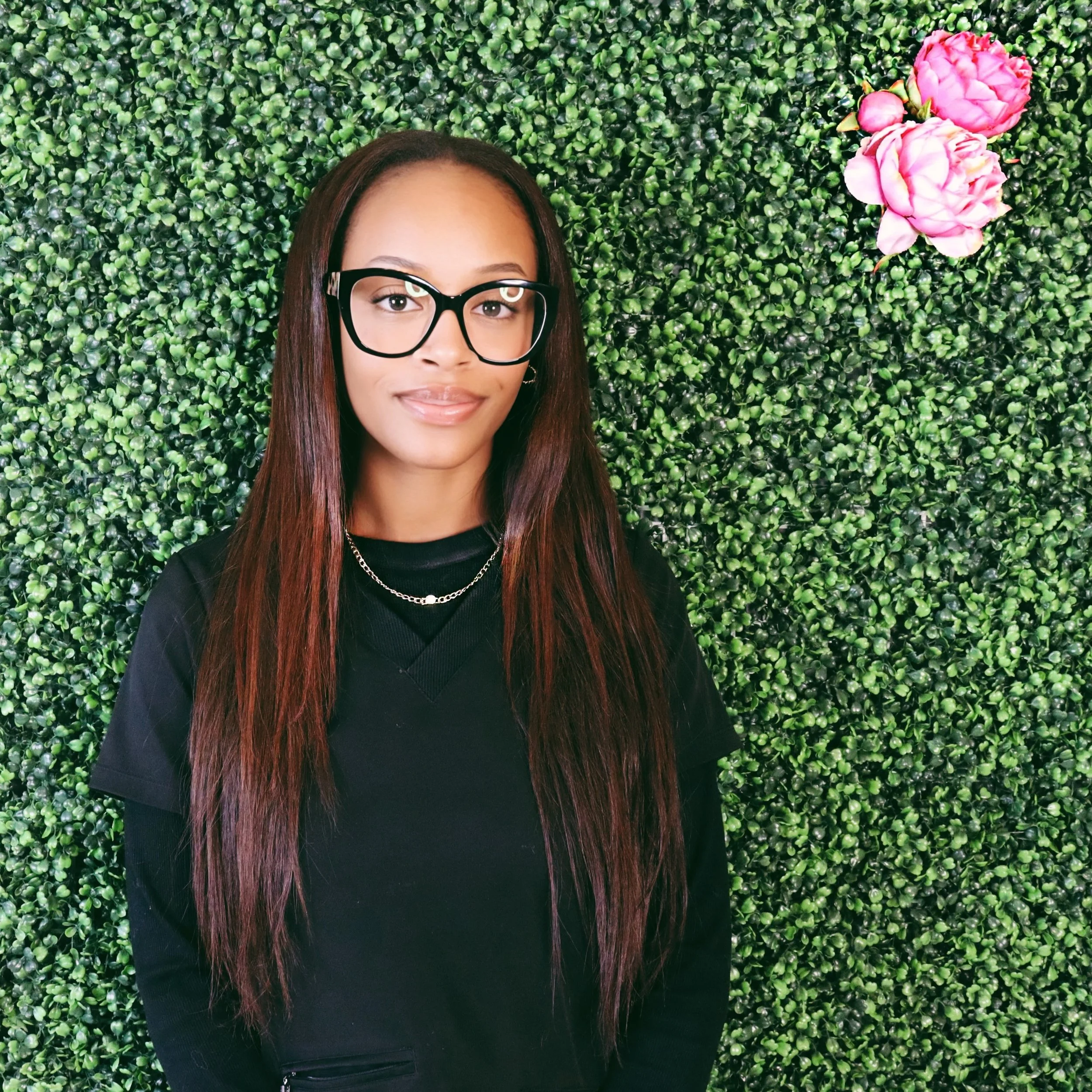Young woman with long straight hair, glasses, and wearing a black top standing in front of a green leafy wall with pink flowers.