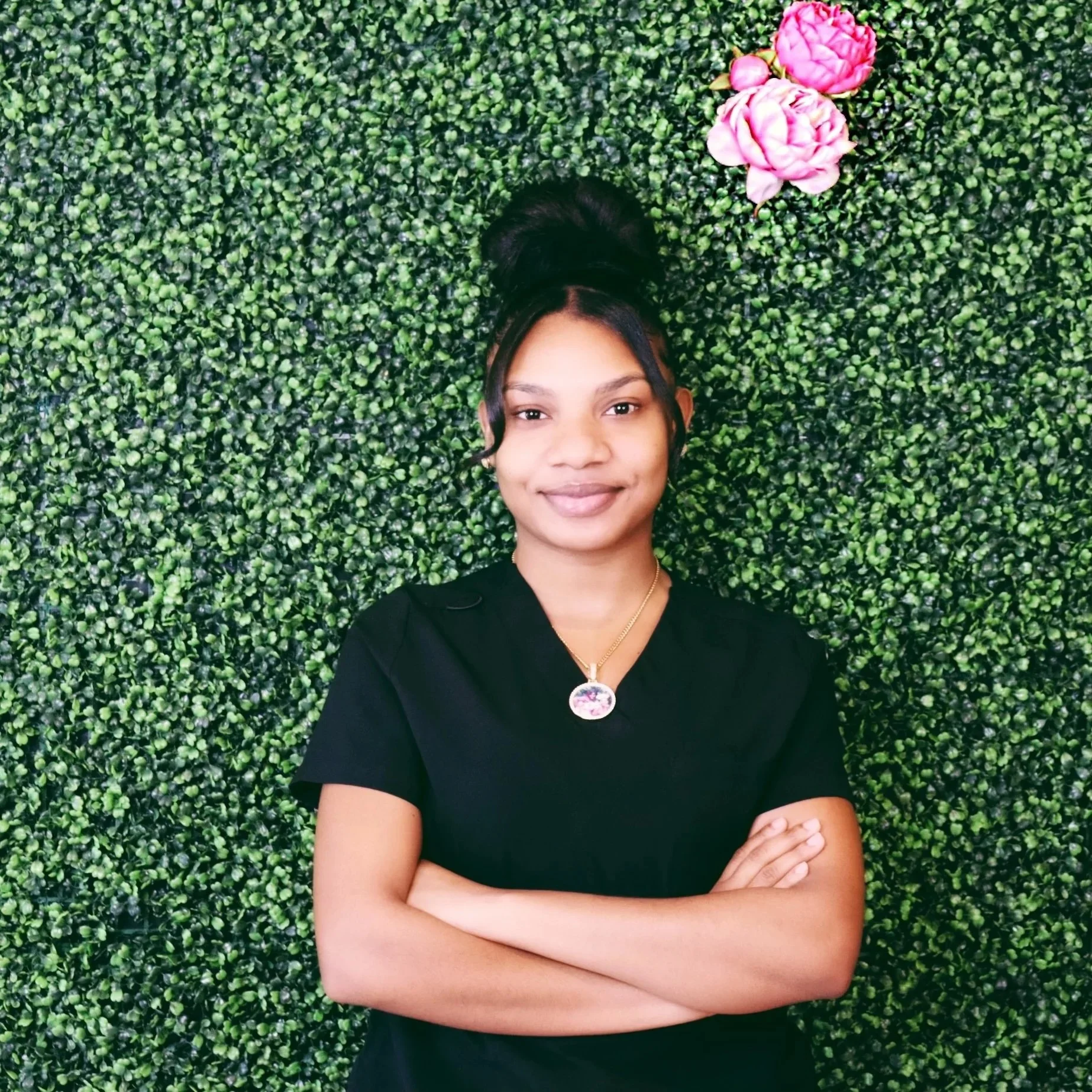 A smiling woman with arms crossed, wearing a black shirt and necklace, standing in front of a green leafy wall with pink flowers.