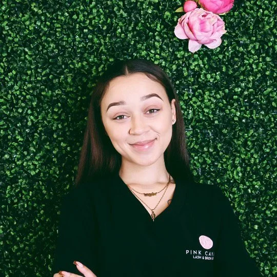 Young woman smiling with crossed arms in front of green hedge wall with pink roses, wearing black top and layered necklaces.