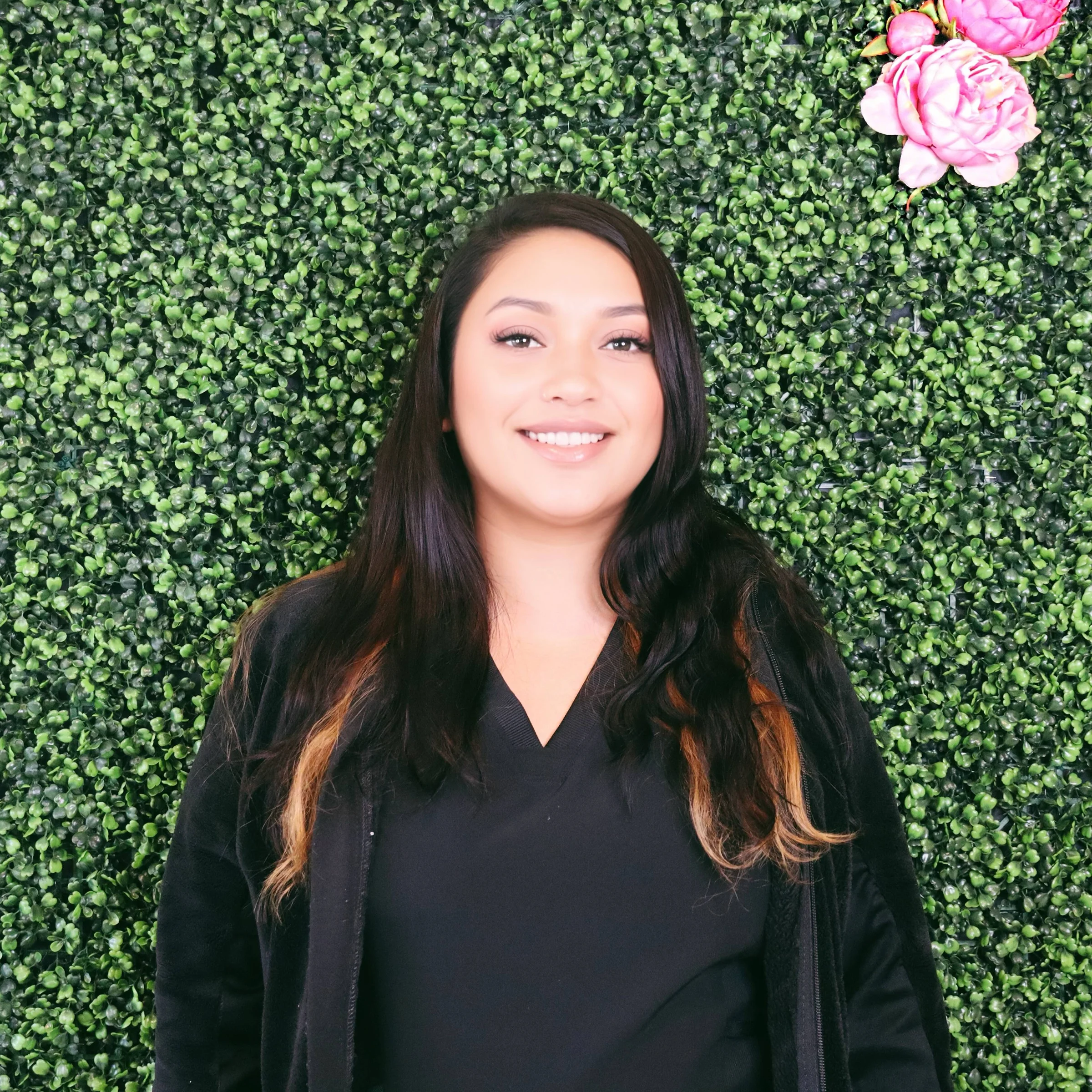 Smiling woman with long dark hair standing in front of a green leafy hedge with pink flowers.