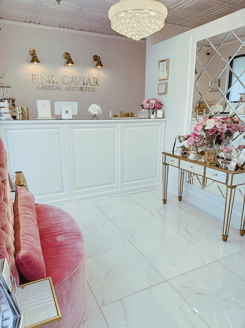 Reception area of Pink Caviar Medical Aesthetics with a white counter, pink flowers, and elegant decor, including a chandelier and mirrors.