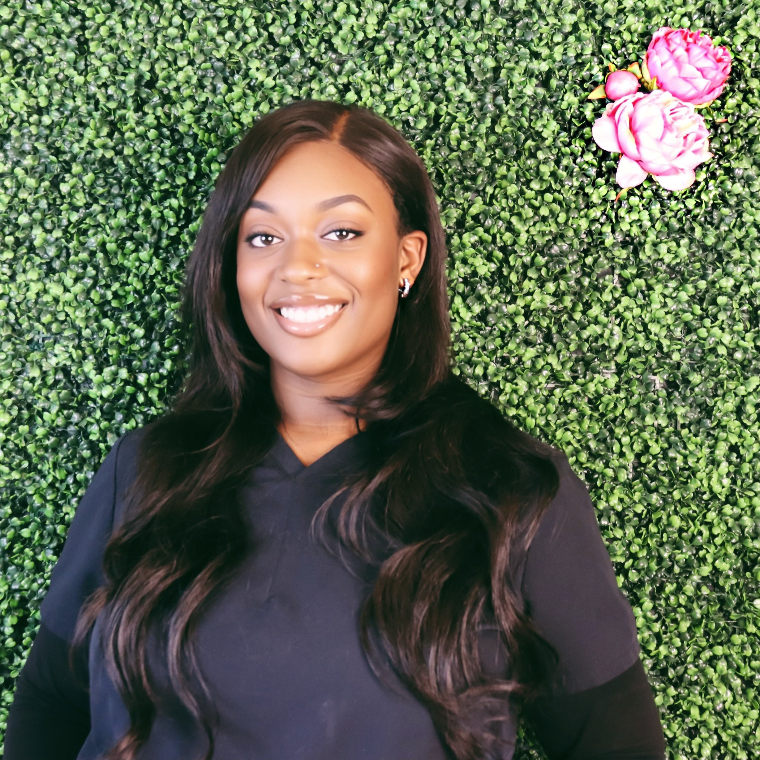 A smiling young woman with long wavy dark hair, wearing a black top, standing in front of a green leafy wall with pink flowers.