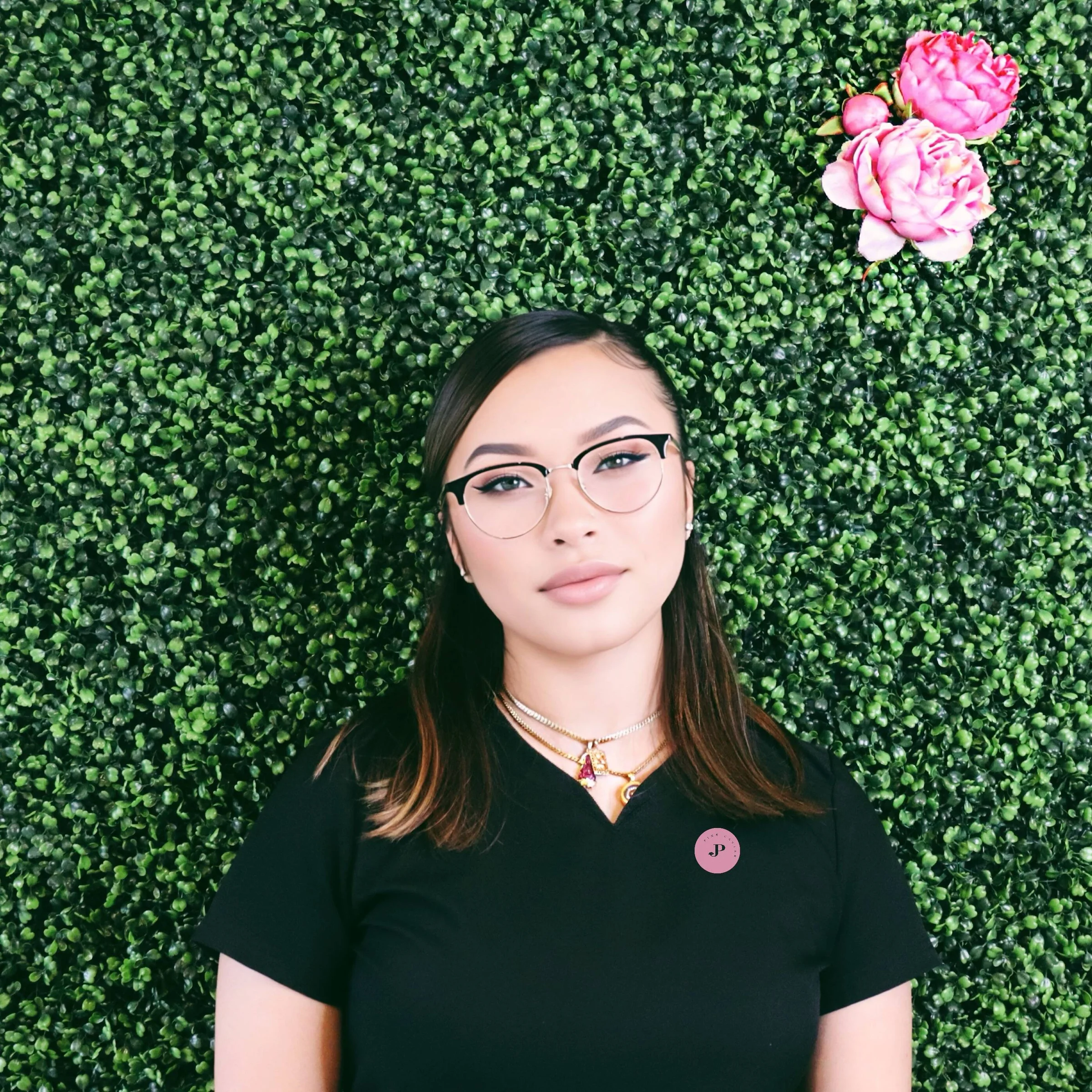 Portrait of a woman with glasses and layered necklaces standing in front of a green leafy wall with pink flowers.