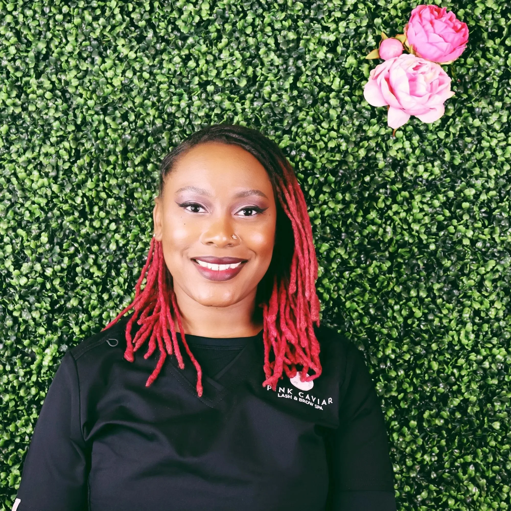 A woman with red dreadlocks smiling in front of a green leafy wall with pink flowers in the top right corner.