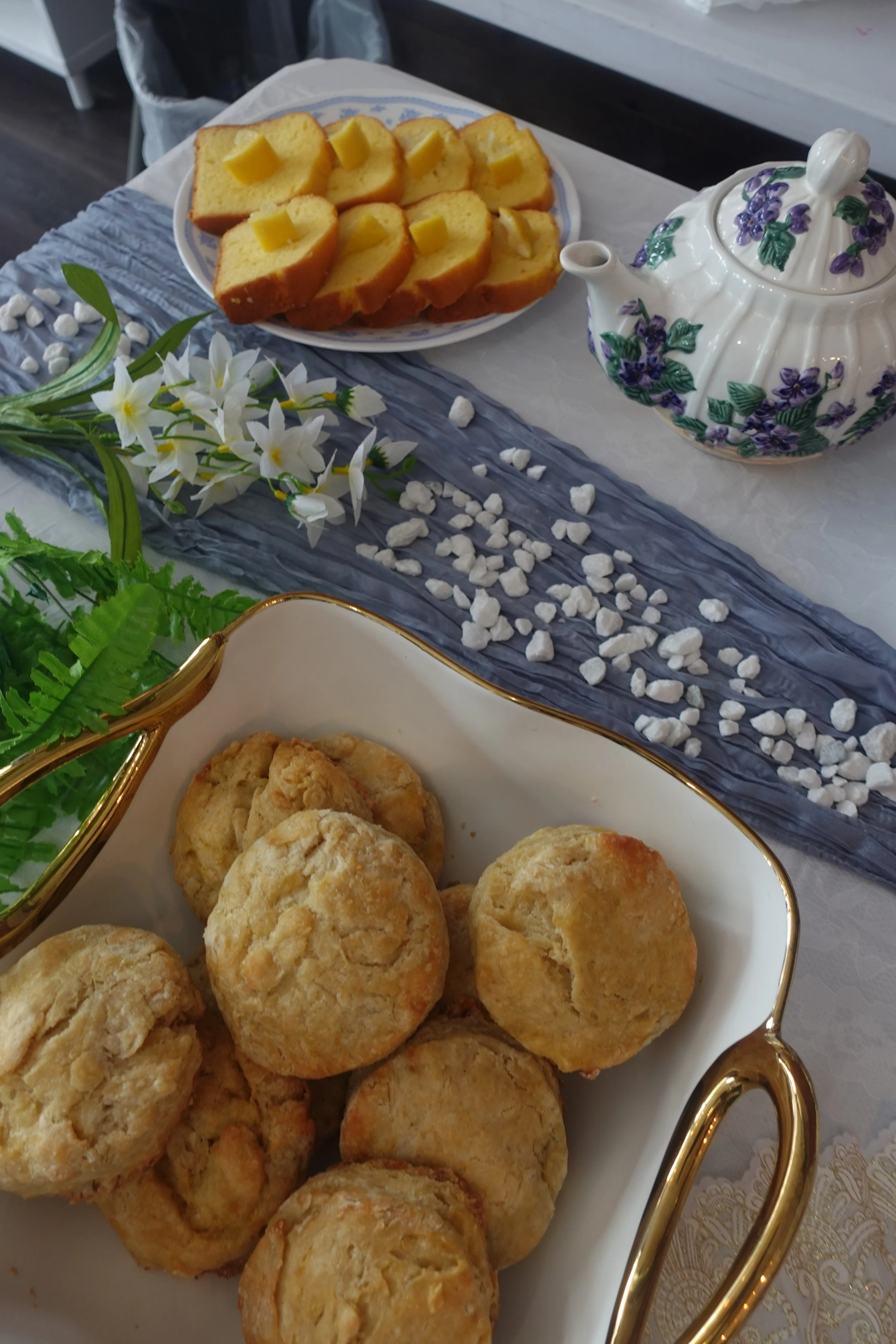 Plate of sliced pound cake with pieces of butter on top, placed on a table with a decorative table runner and a white floral arrangement nearby. A white teapot with purple and green floral design is also on the table.