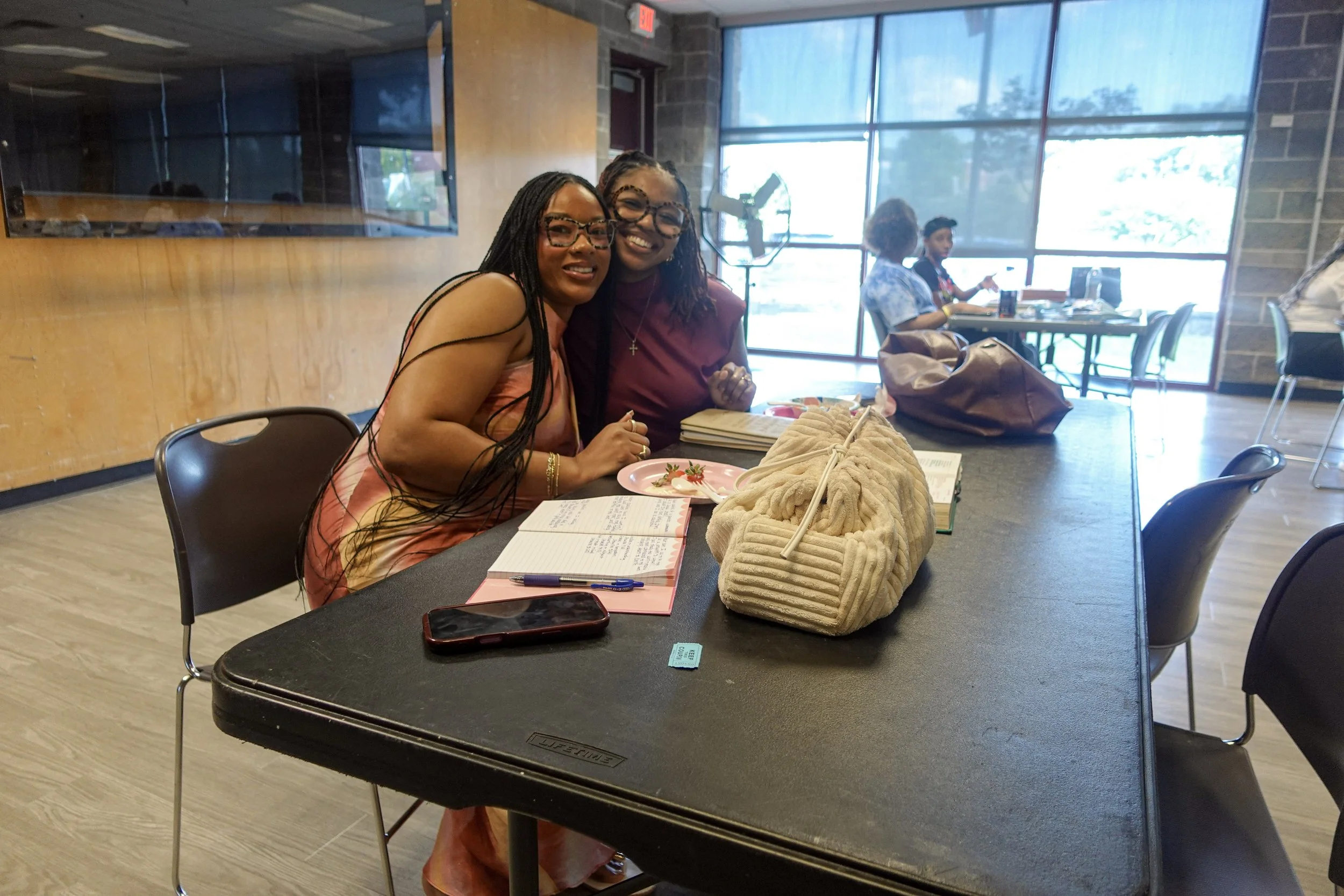 Two women smiling and sitting at a table in a cafe or restaurant, with open notebooks and a beige purse on the table. The background shows large windows and two other women working at a table.