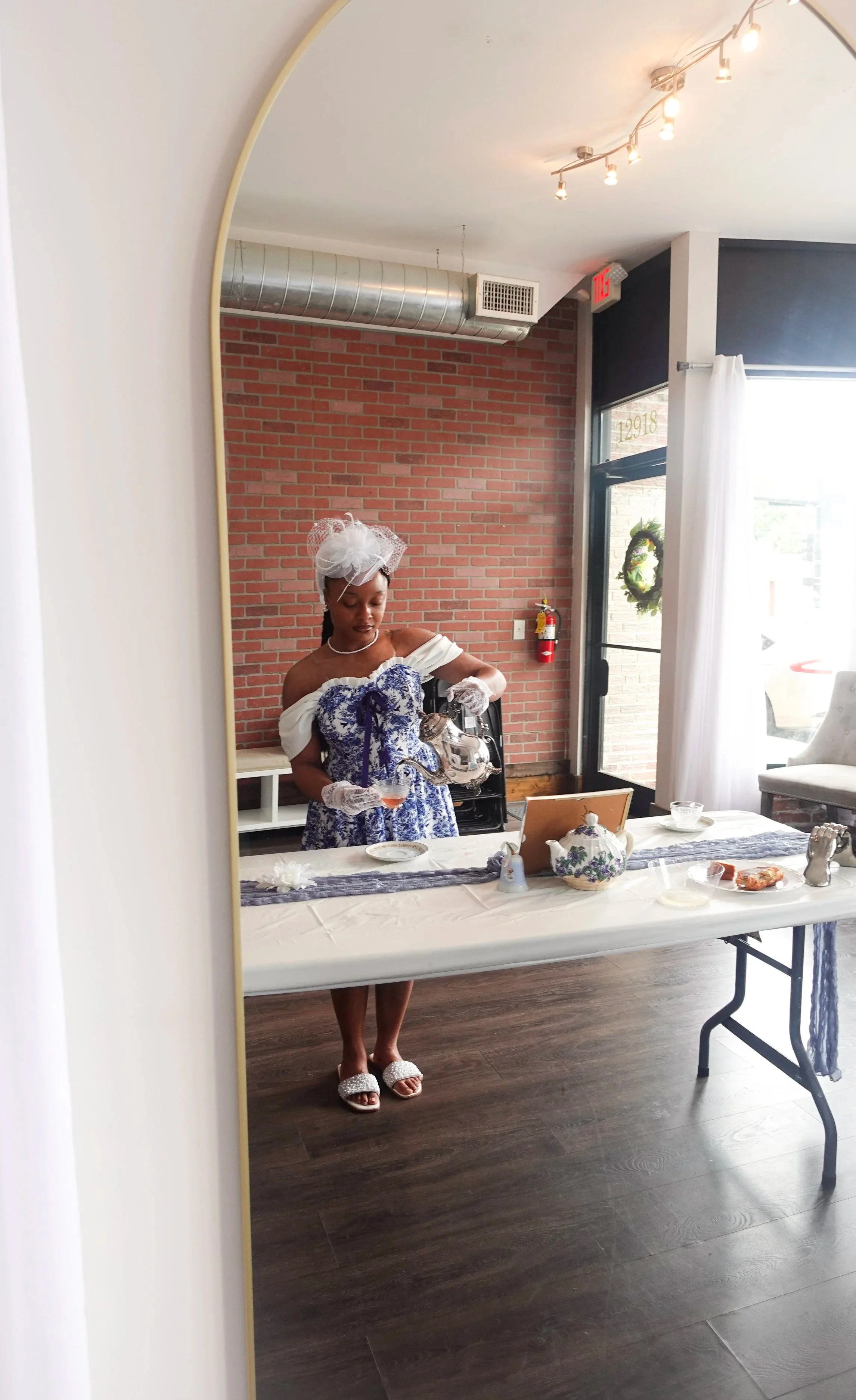 A woman dressed in vintage-style clothing with a white hat and gloves, pouring tea at a table set with a teapot, cups, and plates, seen through a mirror in a room with brick walls and wooden floors, decorated for a celebration or event.