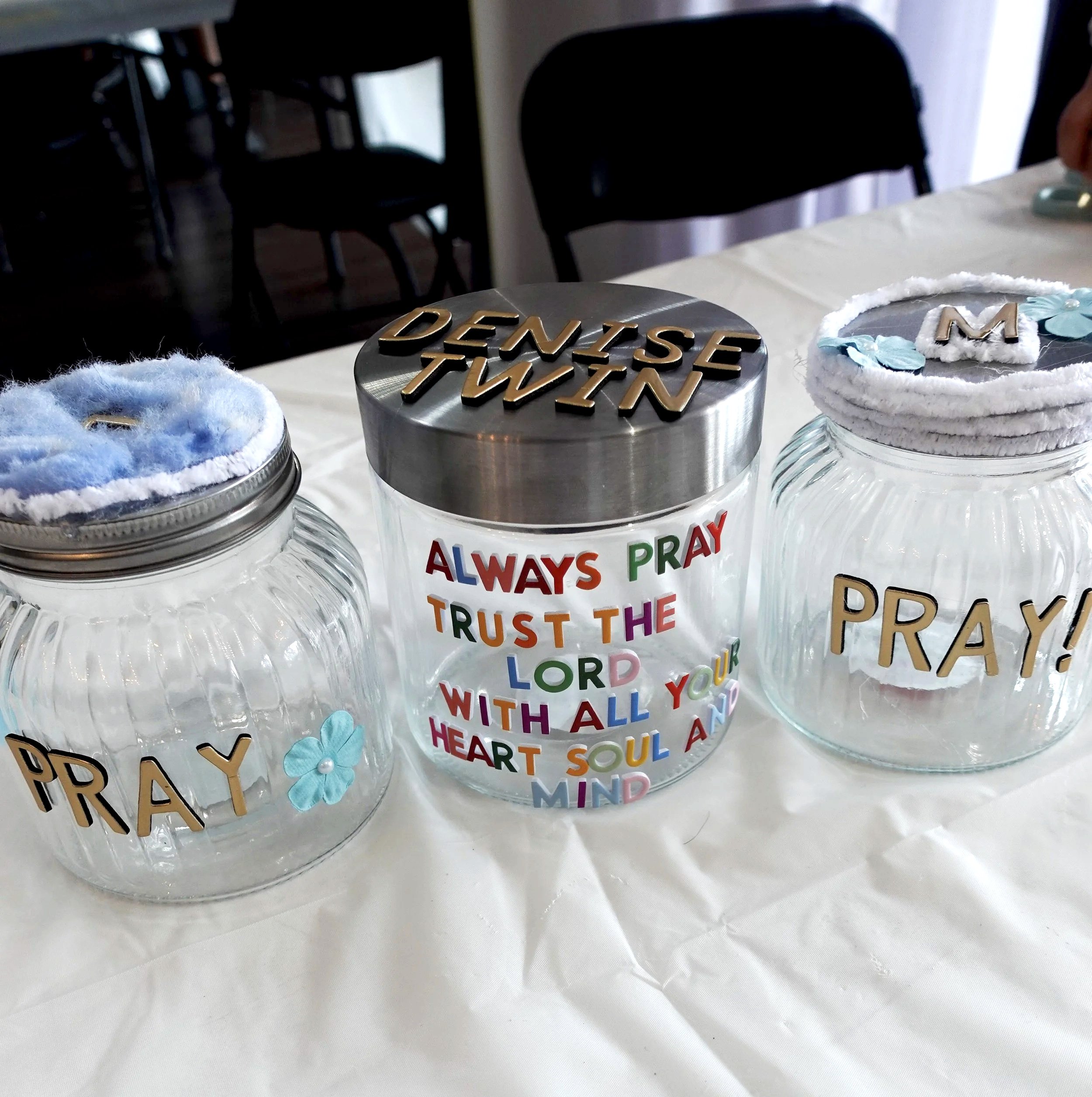 Decorative jars with cotton or felt wreaths, one with a gold word 'DAYS' on the lid, and the other with a colorful phrase 'Always Pray Trust the Lord' on the jar body, placed on a table.