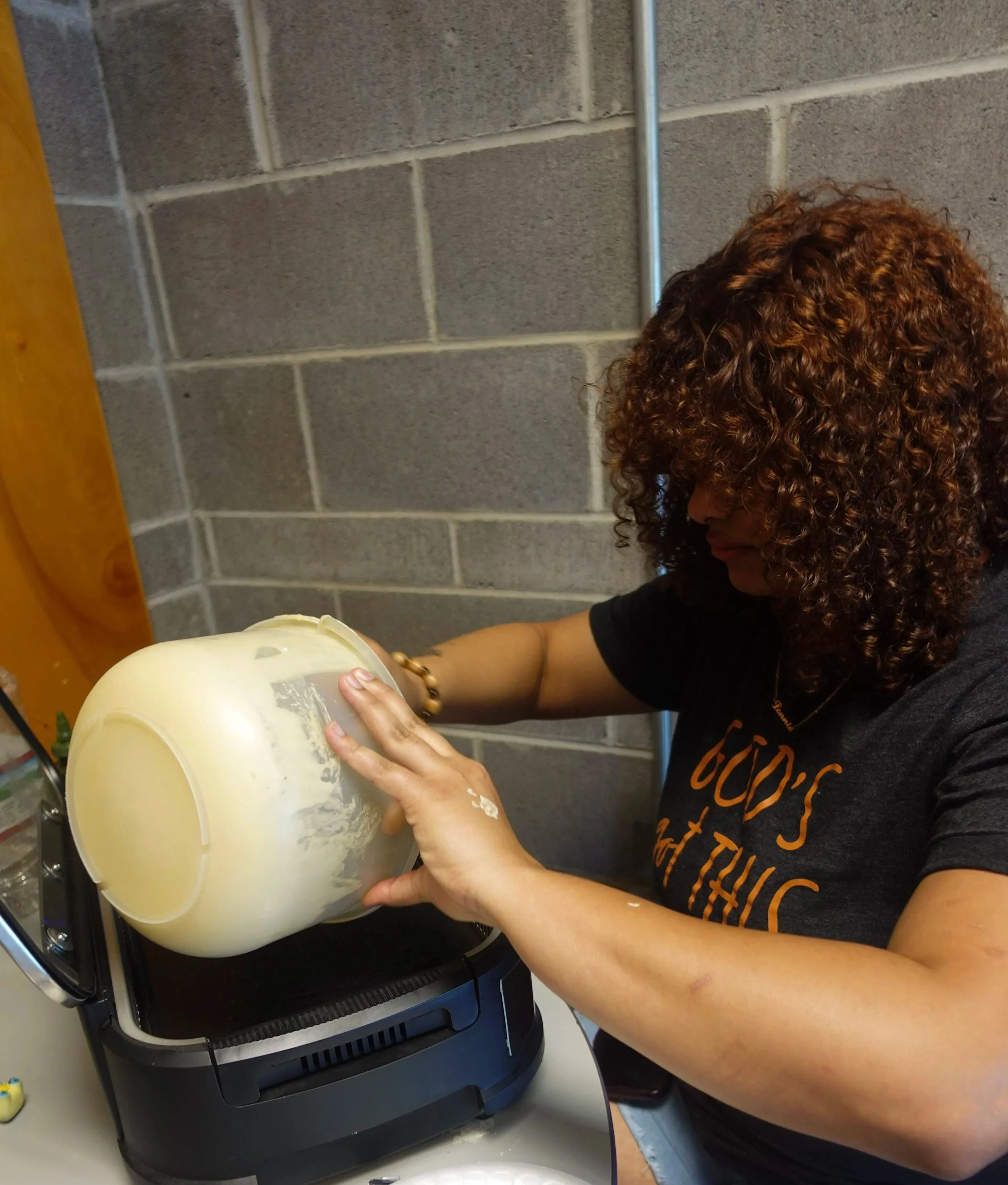 Person with curly hair wearing a black shirt working on a pumpkin with carving tools in a kitchen with gray tiled wall.
