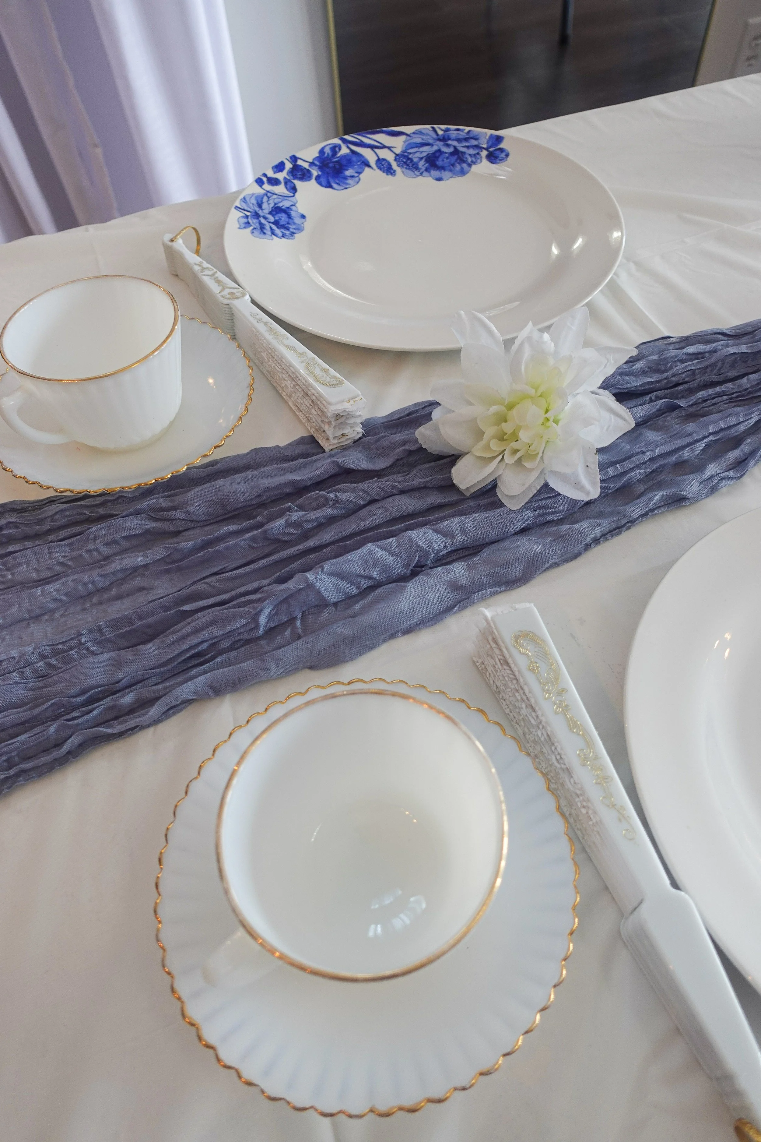 Elegant table setting with a white plate decorated with a blue floral pattern, a matching cup and saucer with gold trim, a white napkin with gold accents, and a white flower with green leaves on a blue textured table runner.