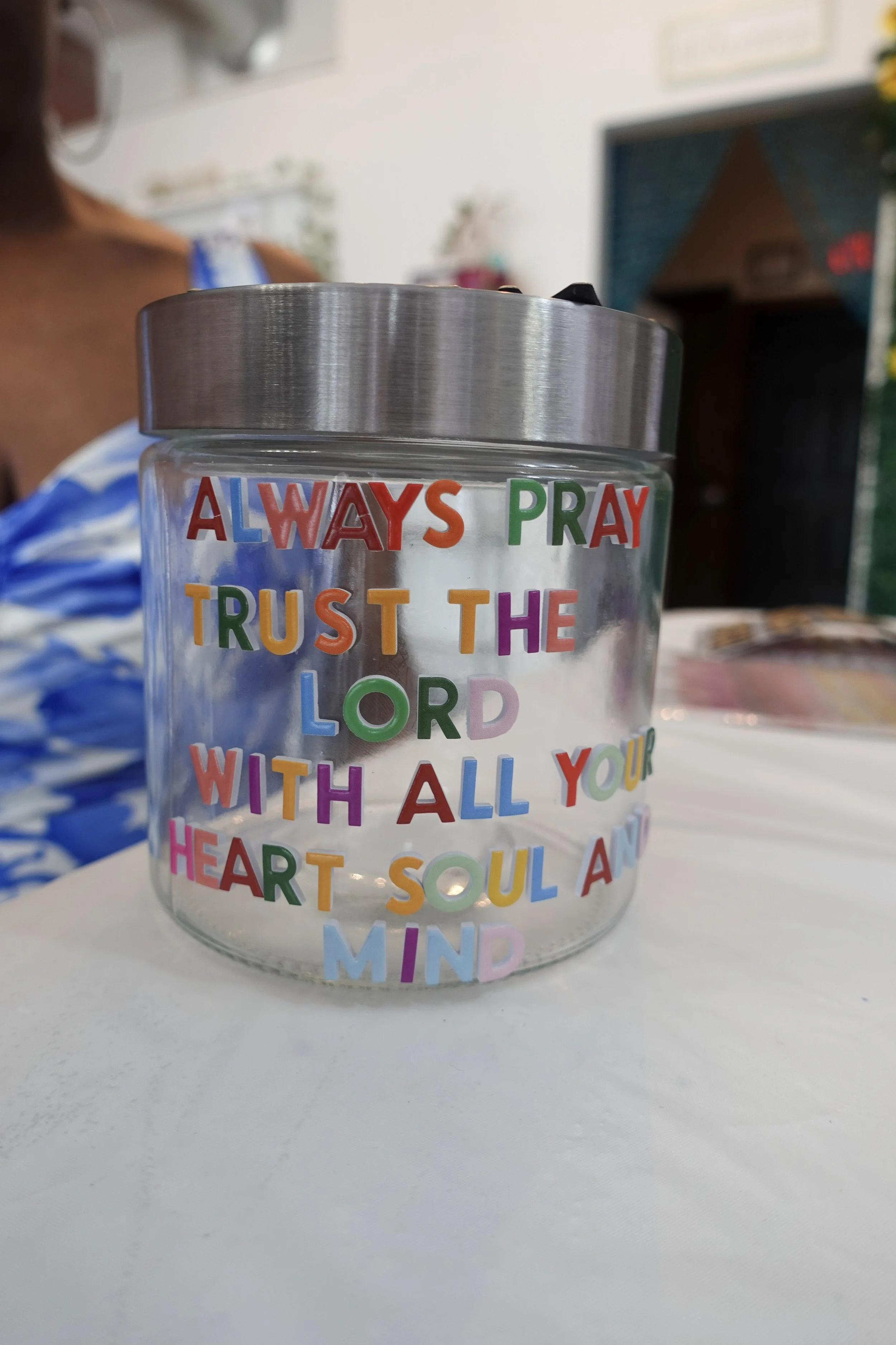 Close-up of a transparent container with colorful text that reads: "ALWAYS PRAY TRUST THE LORD WITH ALL YOUR HEART SOUL AND MIND". Background includes a person wearing a blue and white striped top and a blurry indoor setting.