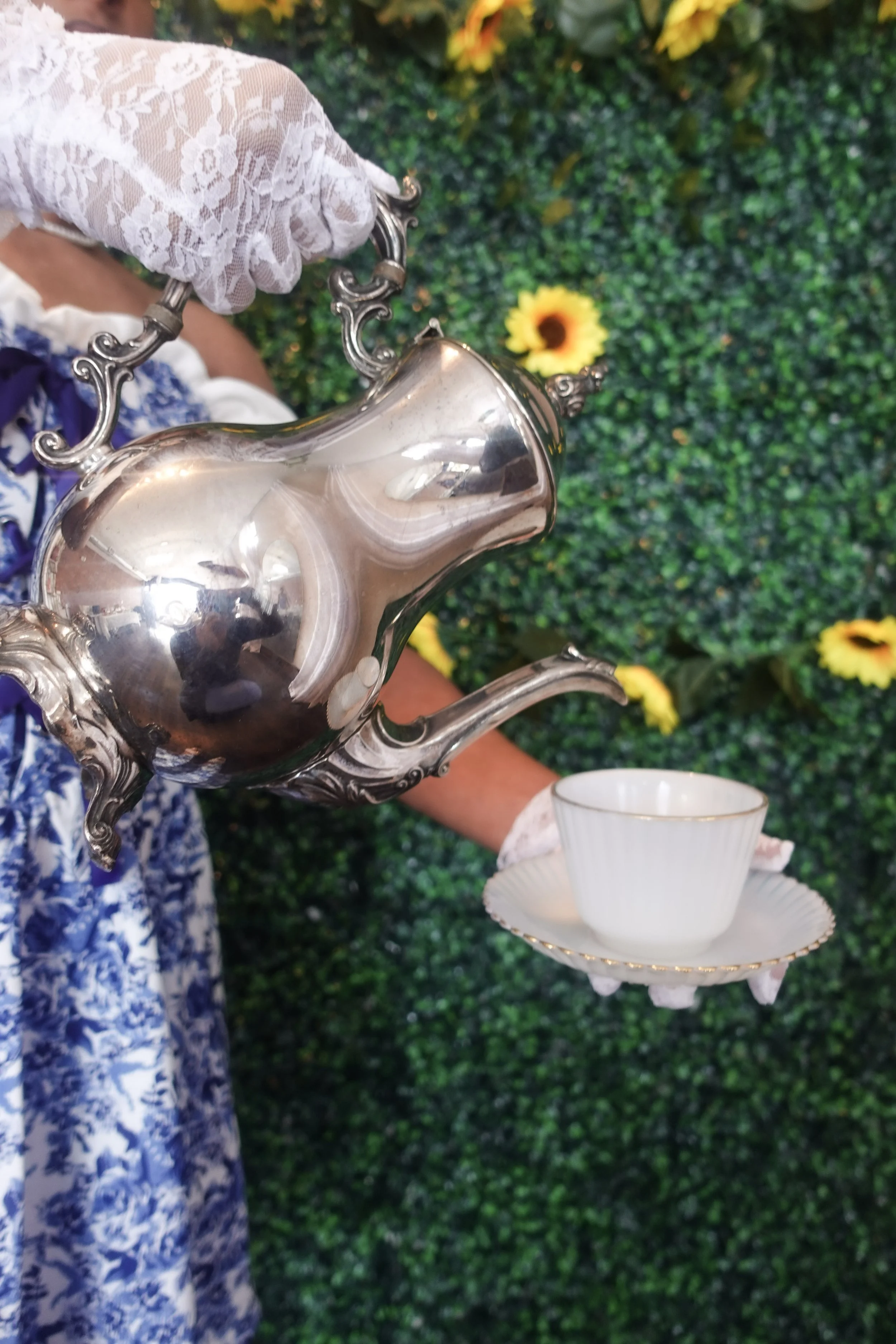 A person wearing a white floral dress and a white lace glove is holding a silver teapot, pouring tea into a white teacup on a matching saucer. The background features green foliage and yellow flowers.
