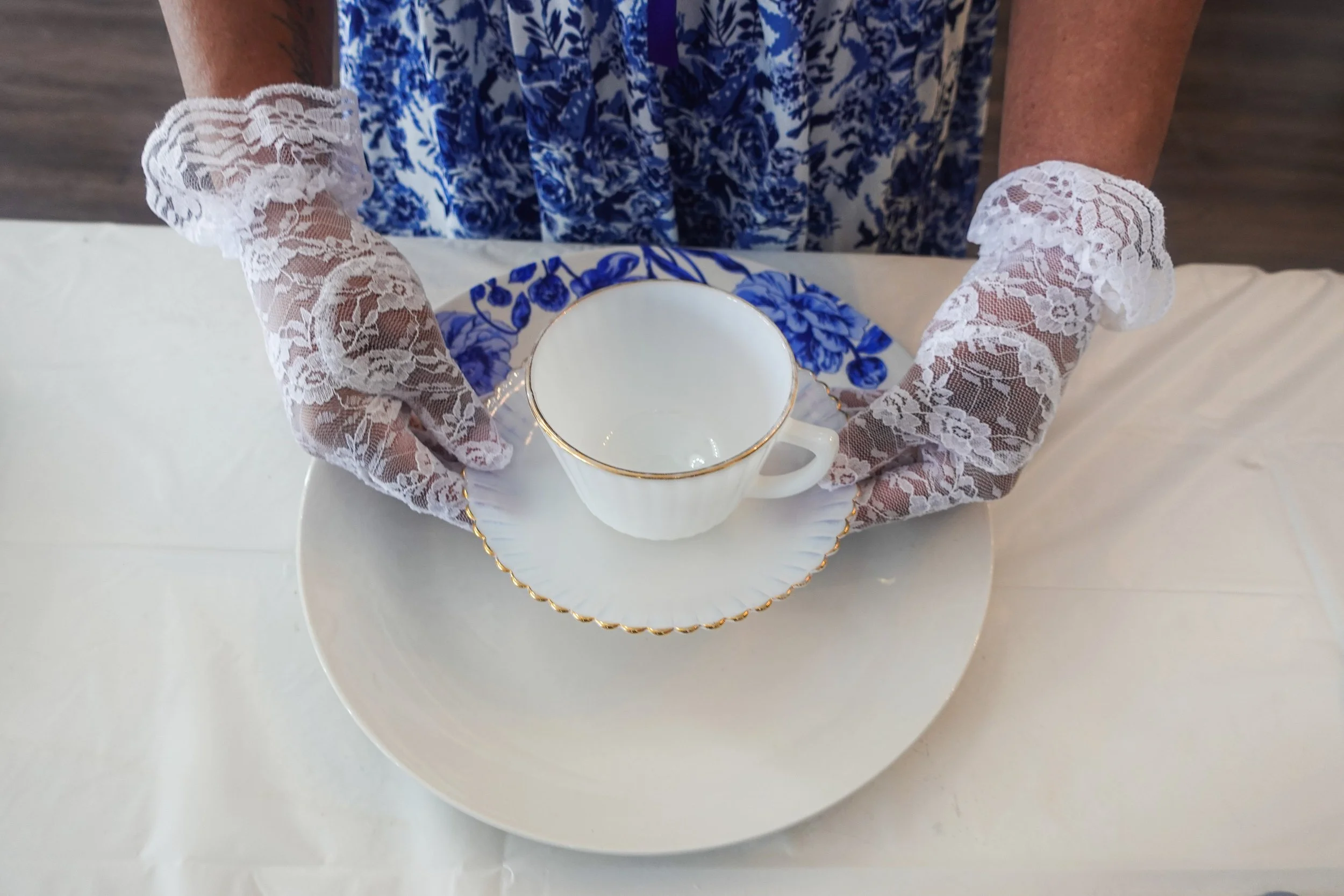 A person wearing lace gloves holding a white and blue floral teacup with a gold rim, placed on a matching saucer on a white table.