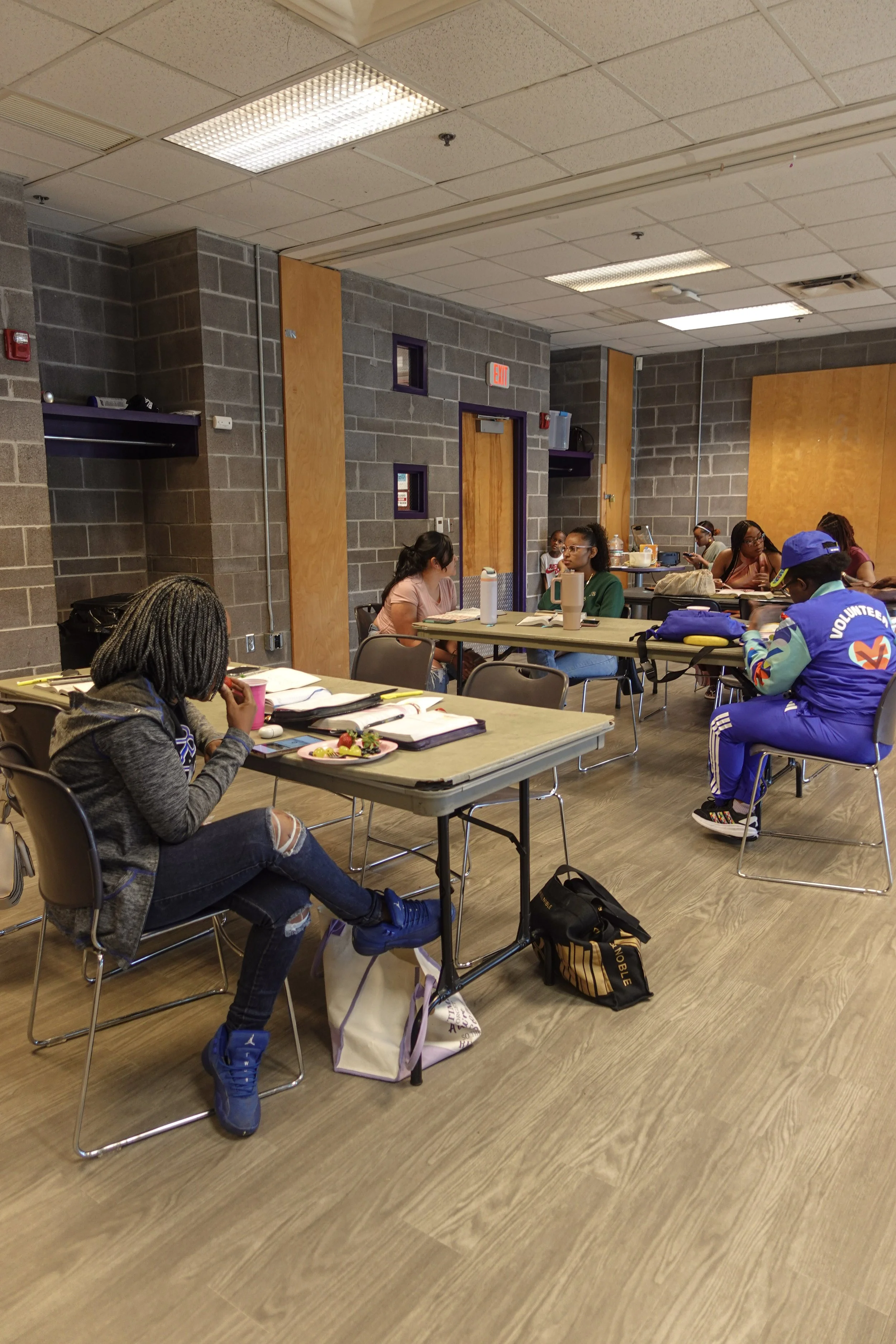 A group of people sitting at tables in a room with gray brick walls and fluorescent lighting, some are engaged in conversation, studying, or eating.
