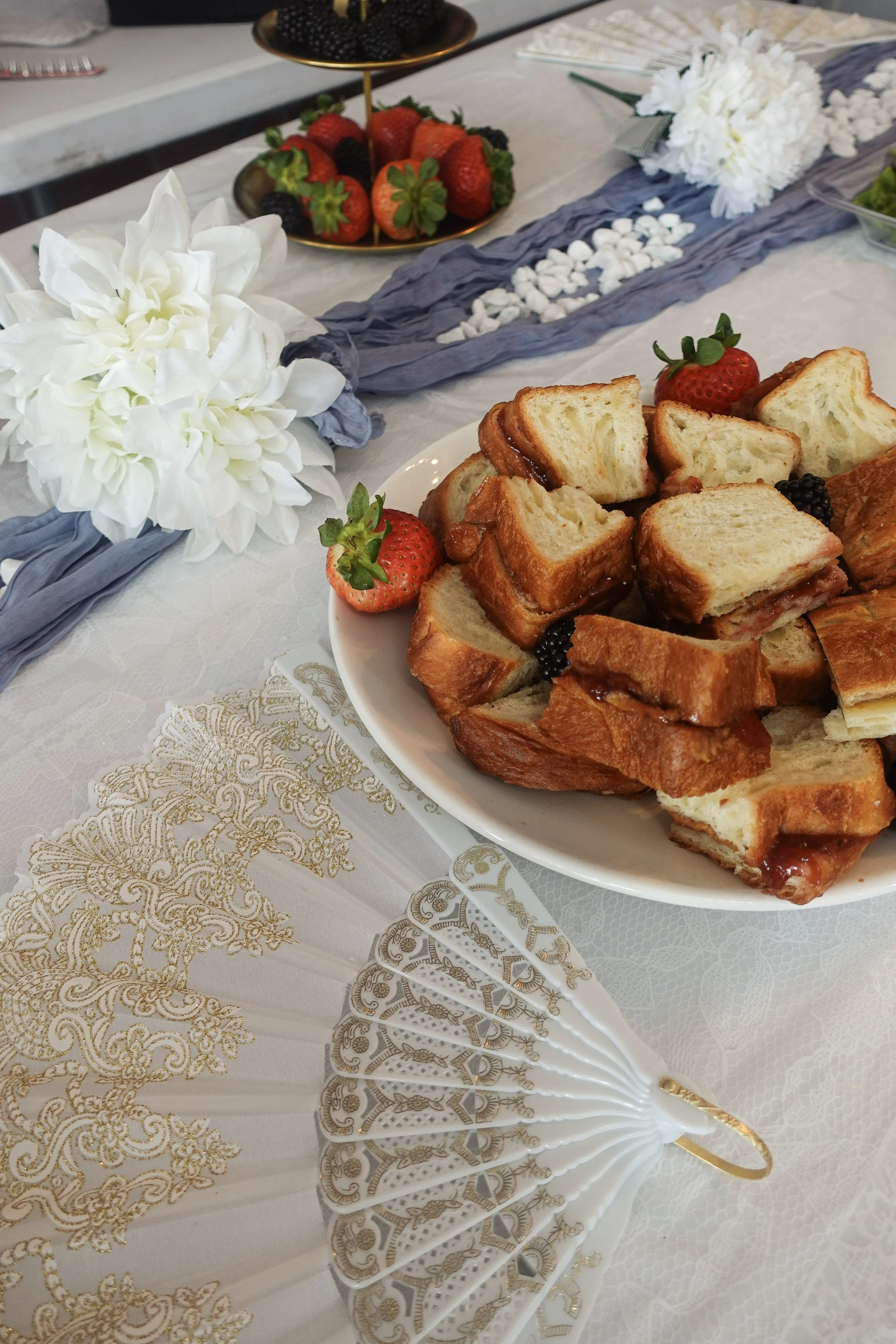 A table setting with a white floral centerpiece, gold and white decorative plates, a bowl of mixed berries, and a plate of sandwiches with strawberries and blackberries. The tablecloth has gold and white lace designs, and there is a blue runner acros