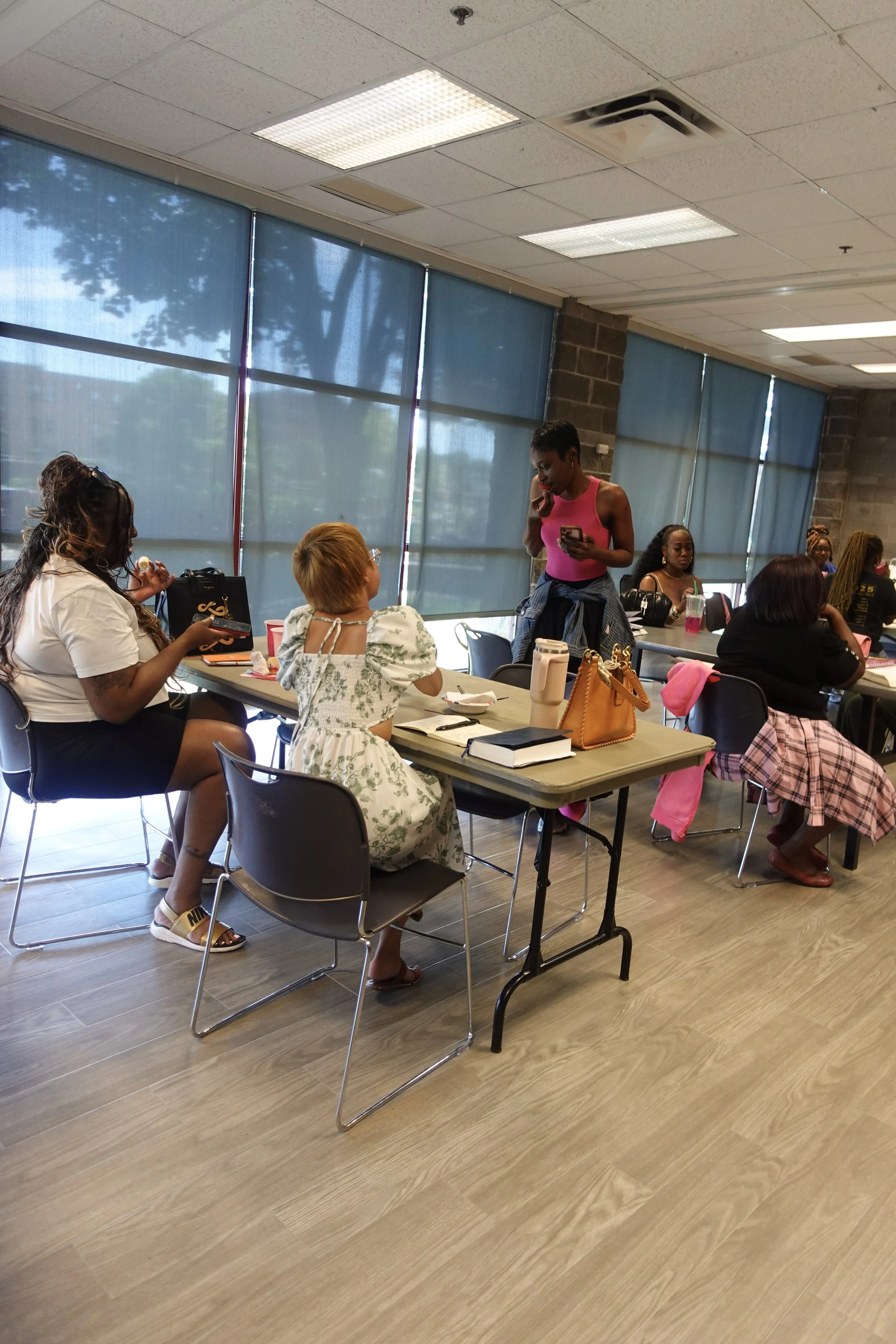 A group of diverse women seated at tables in a meeting room, with some women engaged in conversation and others looking at their phones. The room has large windows with blue blinds and a ceiling with rectangular lights.
