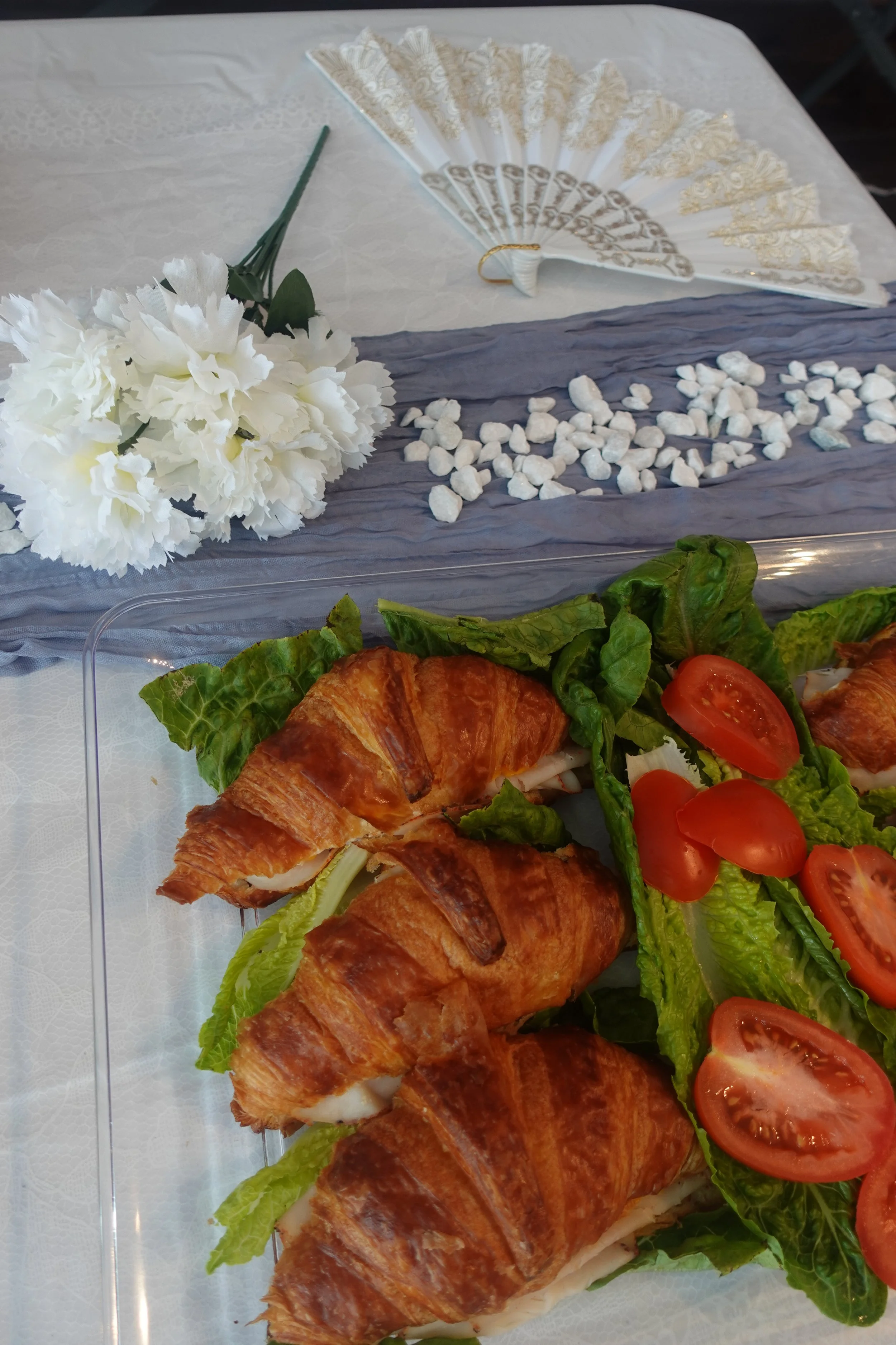 A tray of croissants with lettuce and tomato slices, a flower bouquet, and a decorative fan on a table with a white lace tablecloth.