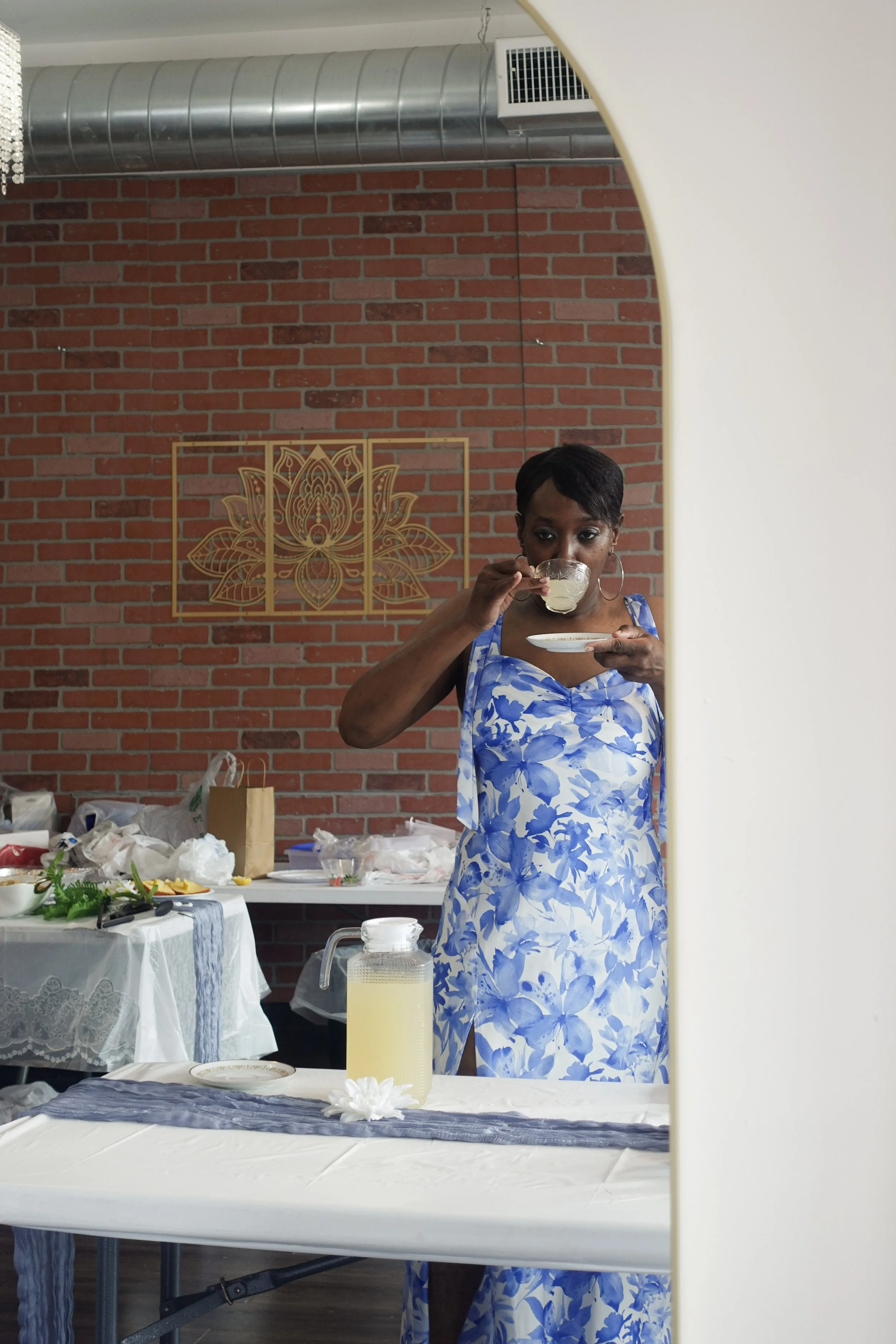Woman in a blue and white floral dress sipping from a cup, reflected in a mirror with a red brick wall and gold decorative wall art behind her.