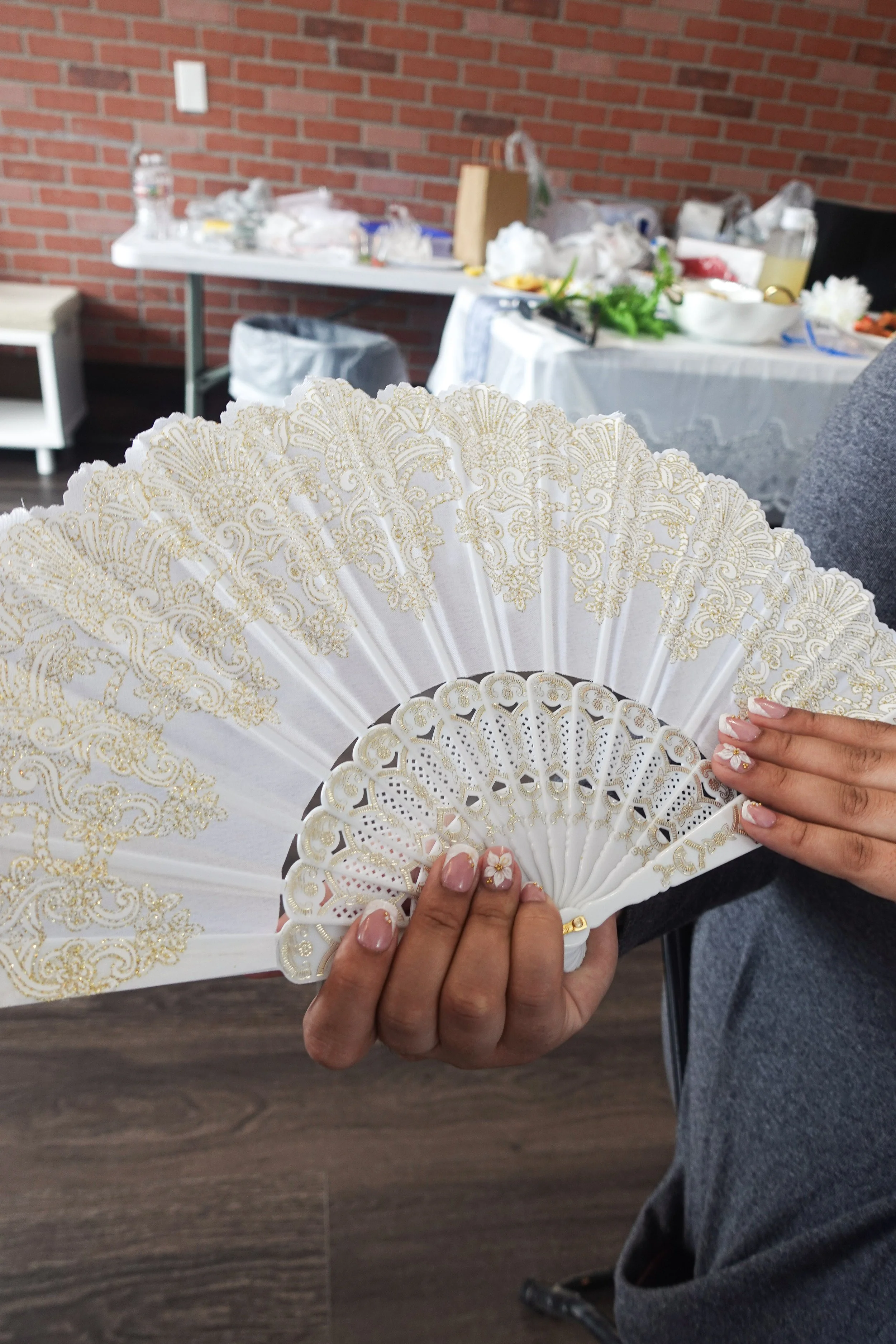 Person holding an ornate white lace hand fan with gold embroidery, with a table and brick wall in the background.