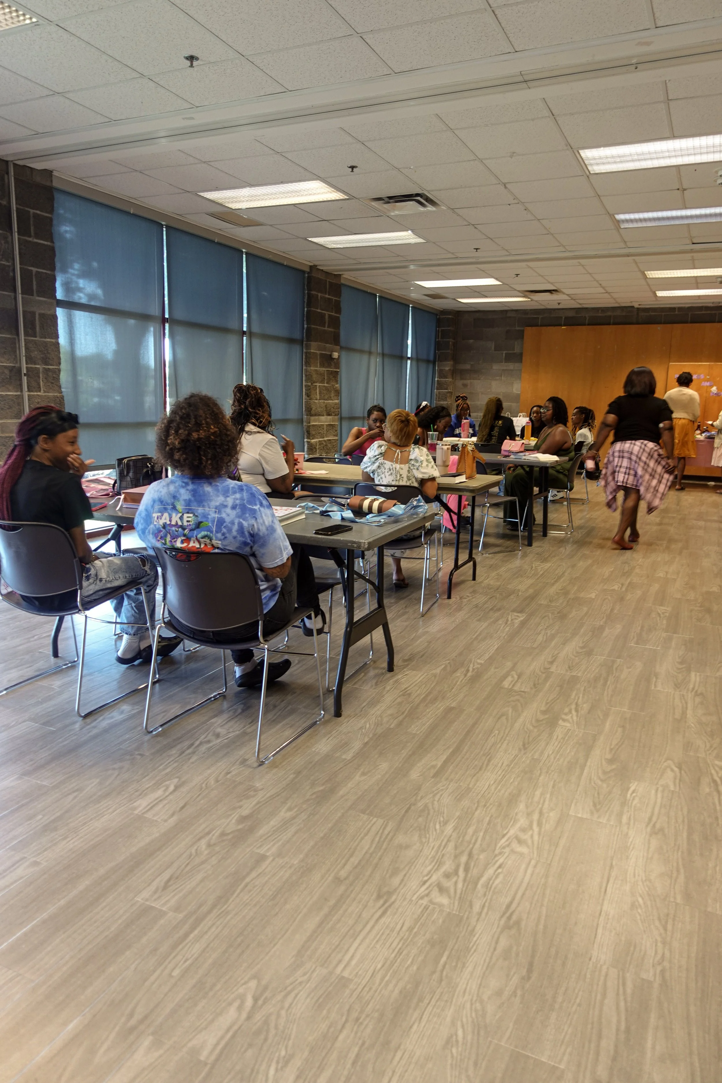 Group of women sitting at tables in a classroom or meeting room with blue window shades and a wooden panel on the wall, some women are smiling and talking, and a woman is walking by.