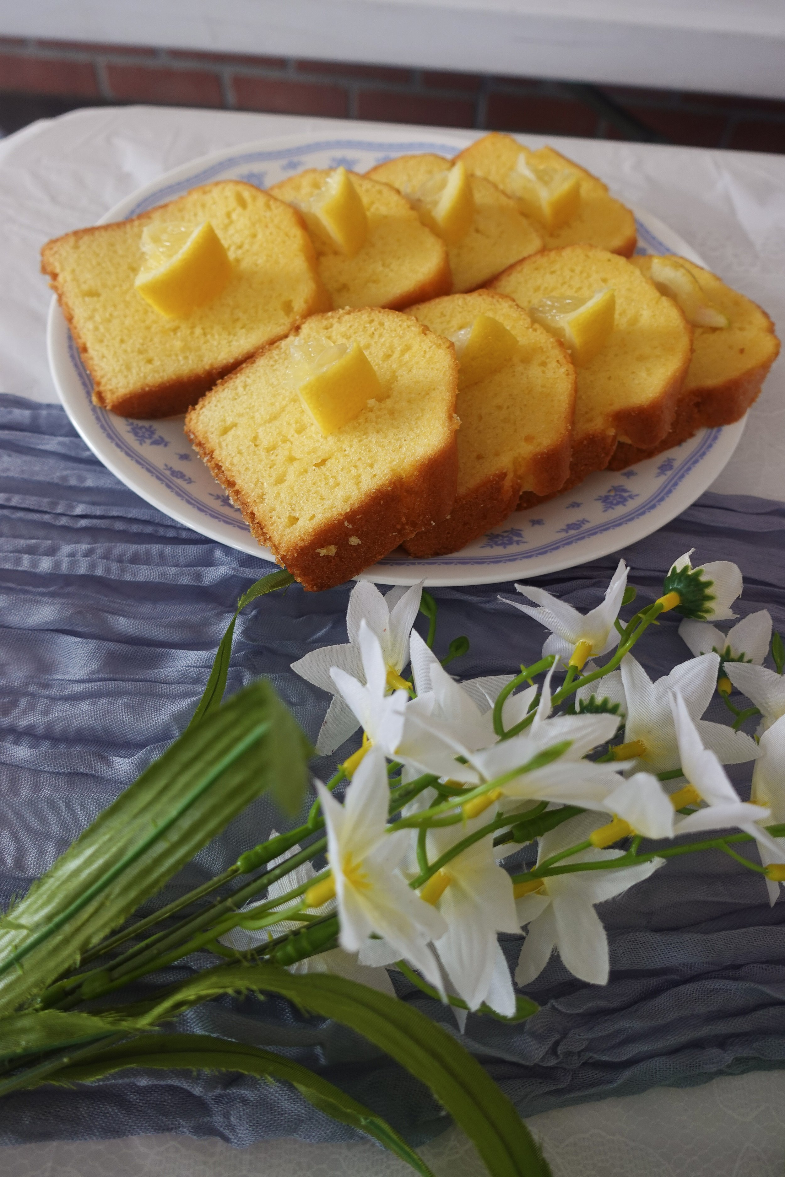 Slices of yellow cake with lemon wedges on top, arranged on a white plate, with white flowers in the foreground.