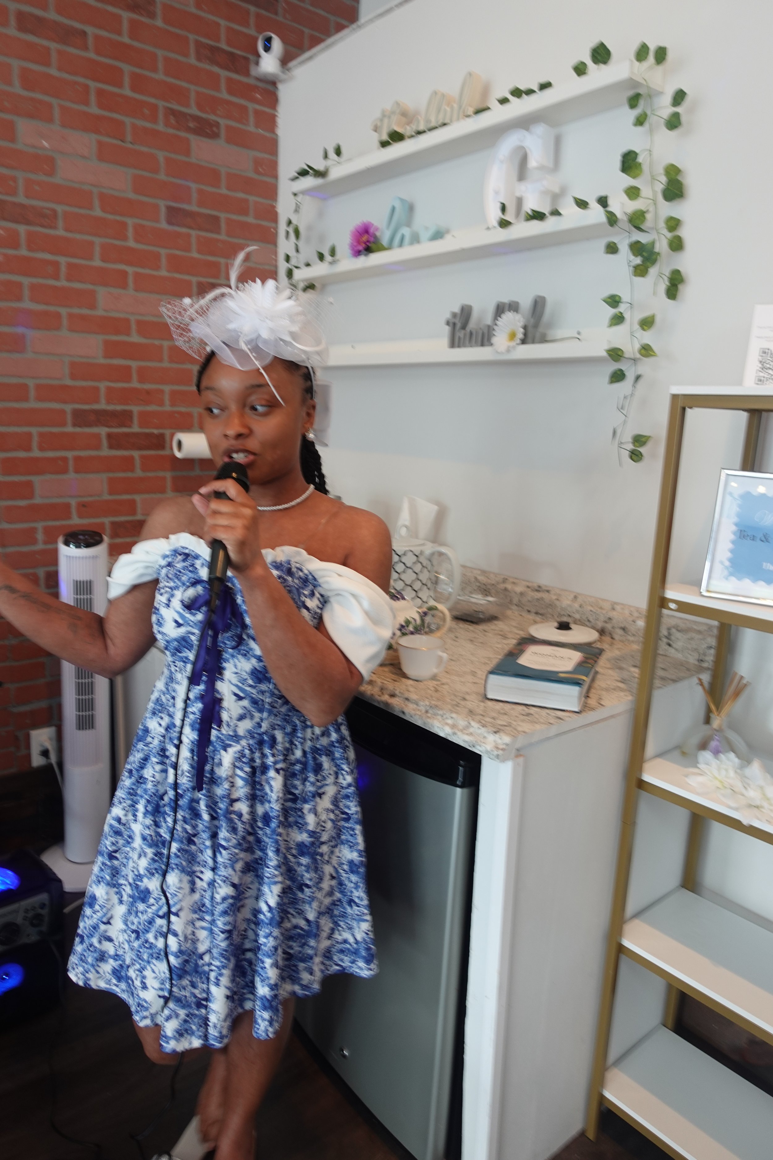 Woman in blue and white dress with a white fascinator hat holding a microphone indoors, with a brick wall and shelves decorated with greenery and decorative items in the background.