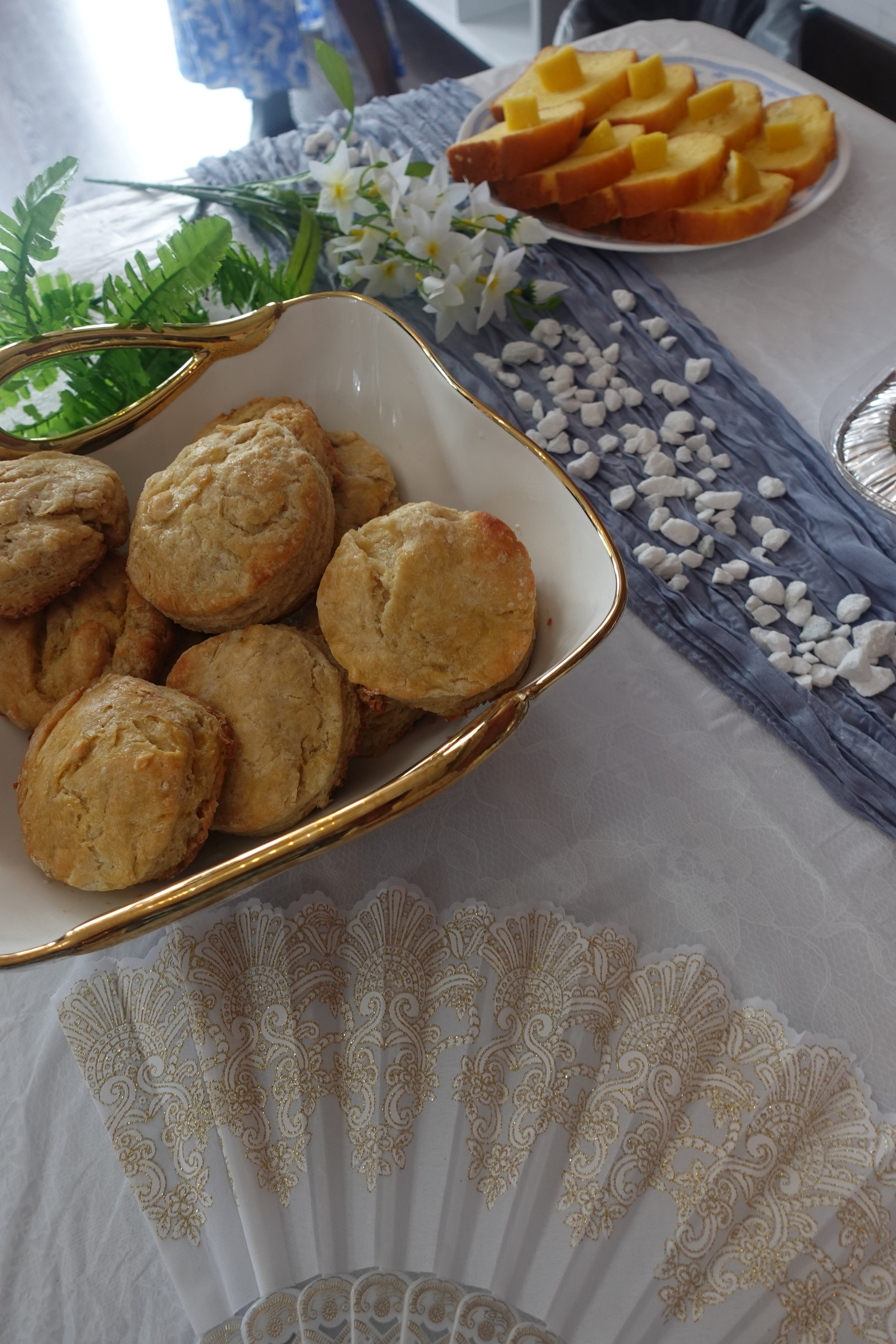 A table setup with a floral and elegant theme, featuring a white and gold decorative paper, a basket of what appears to be cookies or scones, a plate of sliced bread with cheese, and a centerpiece of white flowers and green leaves with scattered whit