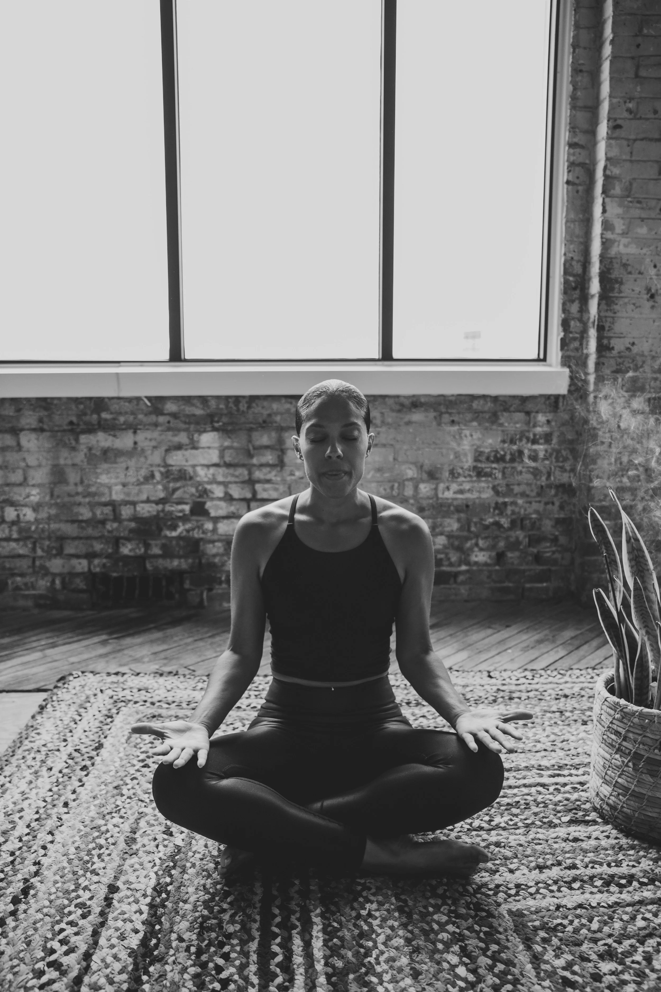Person meditating in a cross-legged position on a patterned rug in a room with brick walls and large windows.