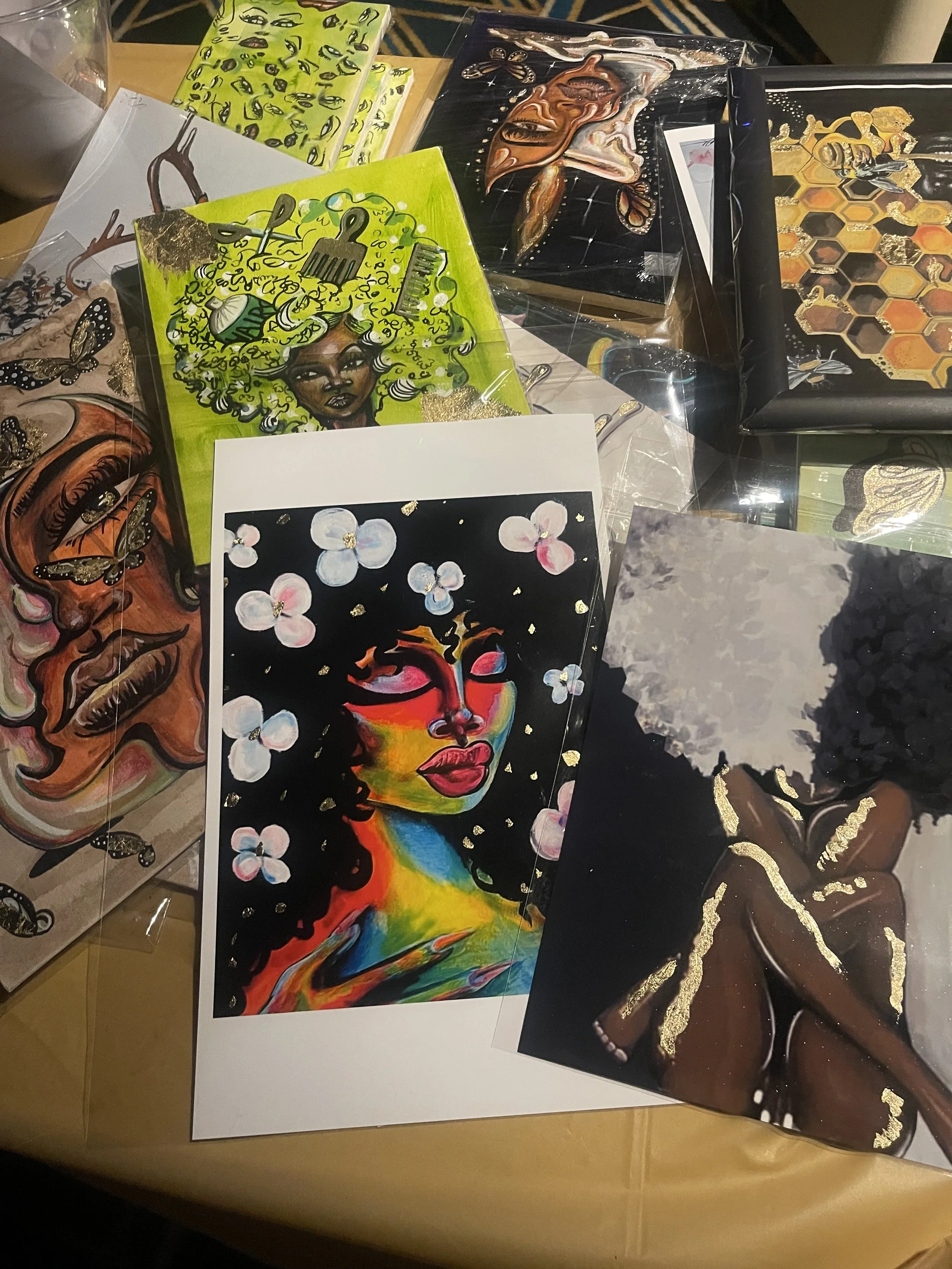 A display of various colorful and artistic paintings and illustrations, including a portrait of a woman with vibrant rainbow-colored hair, an abstract face with geometric accents, and a butterfly design, arranged on a table with some protective plast