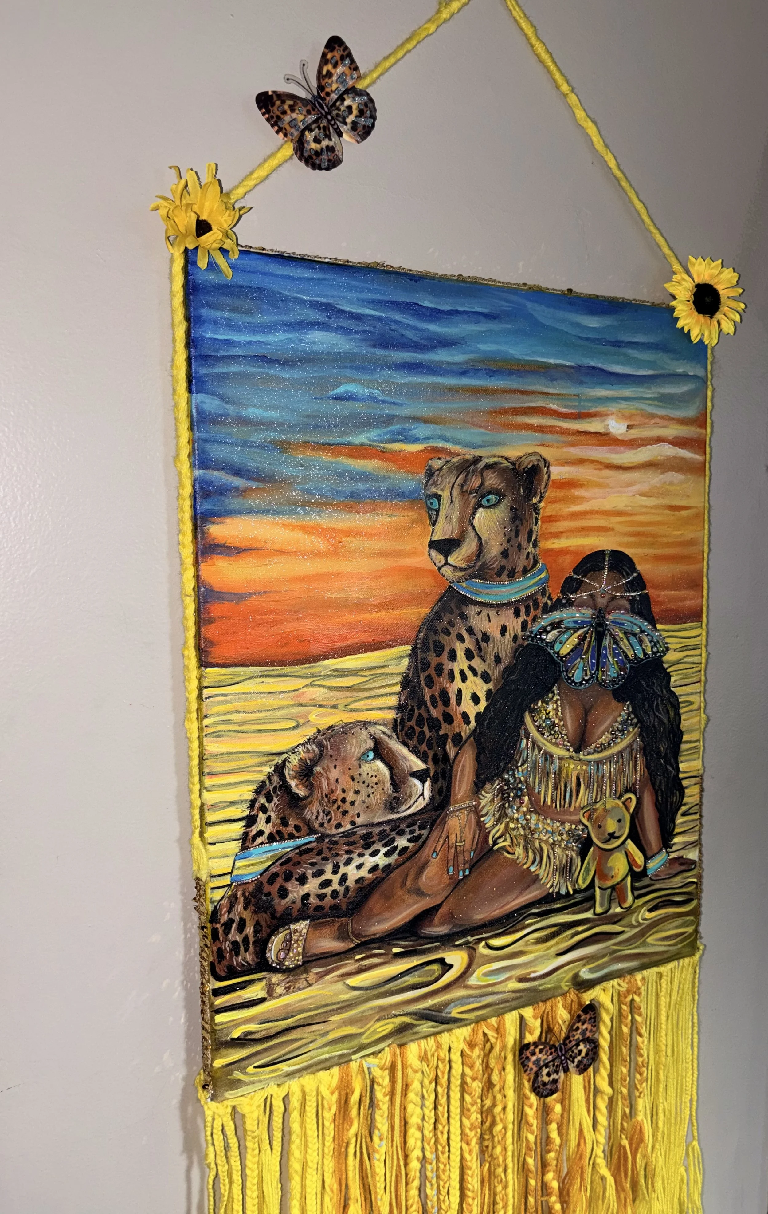 Colorful wall hanging featuring a sunset sky with clouds, a woman with long black hair dressed as an indigenous figure, and three cheetahs. The hanging is decorated with yellow yarn and butterflies, and has yellow flowers at the top corners.