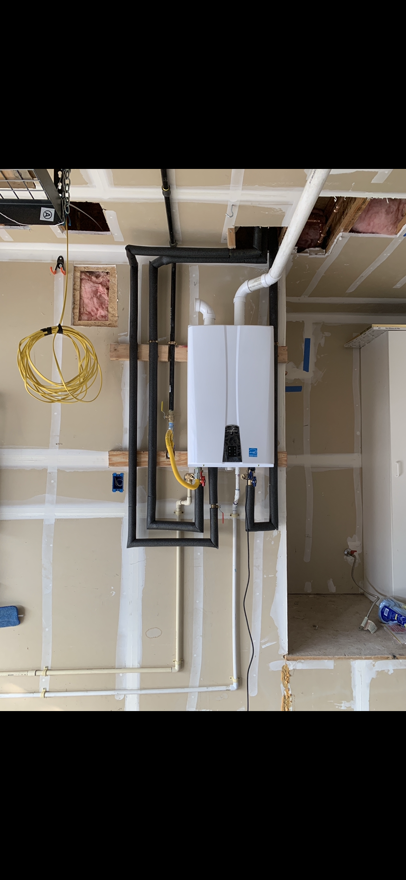 Garage wall with tankless water heater, insulated pipes, and yellow electrical wire.