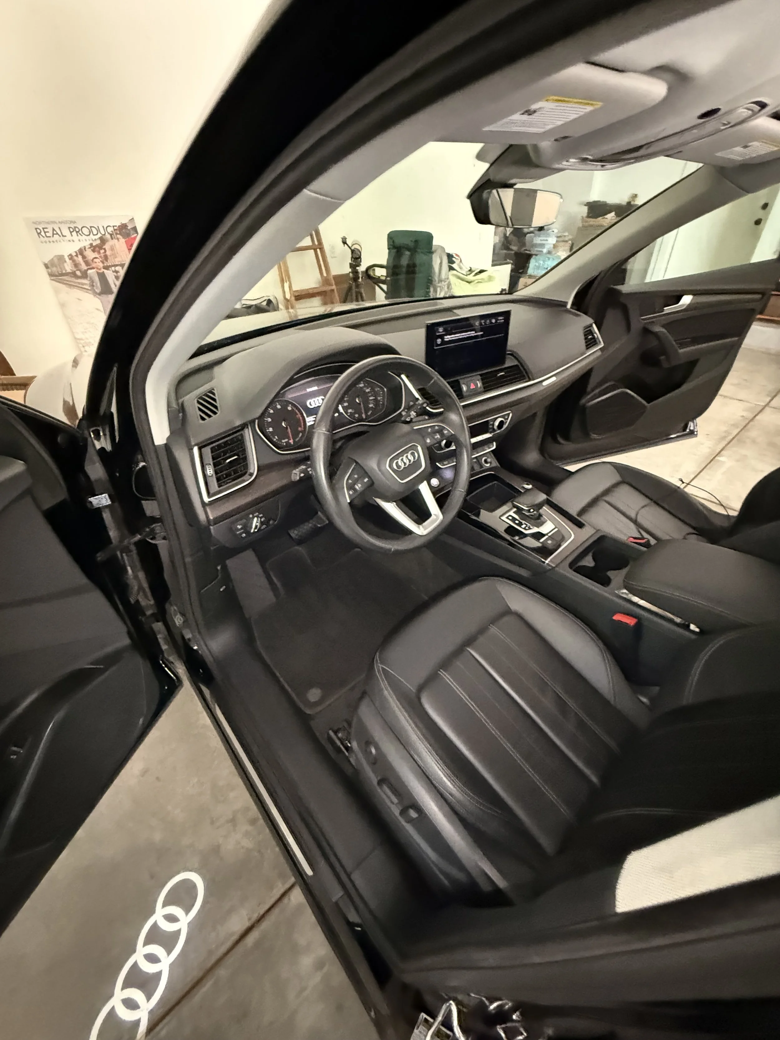 Inside an Audi vehicle showing the dashboard, steering wheel, front seats, and the open driver side door. In the background, there are some storage items and a tripod.