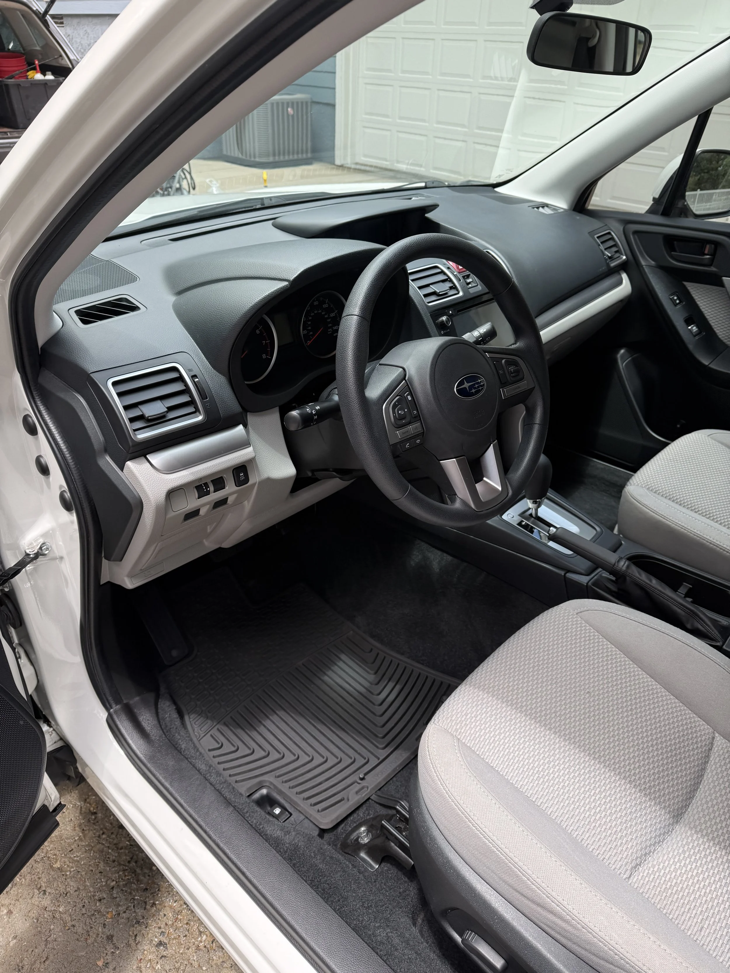 Interior of a car with a manual transmission, fabric seats, and dashboard showing the steering wheel with the Subaru logo, air vents, and control buttons.