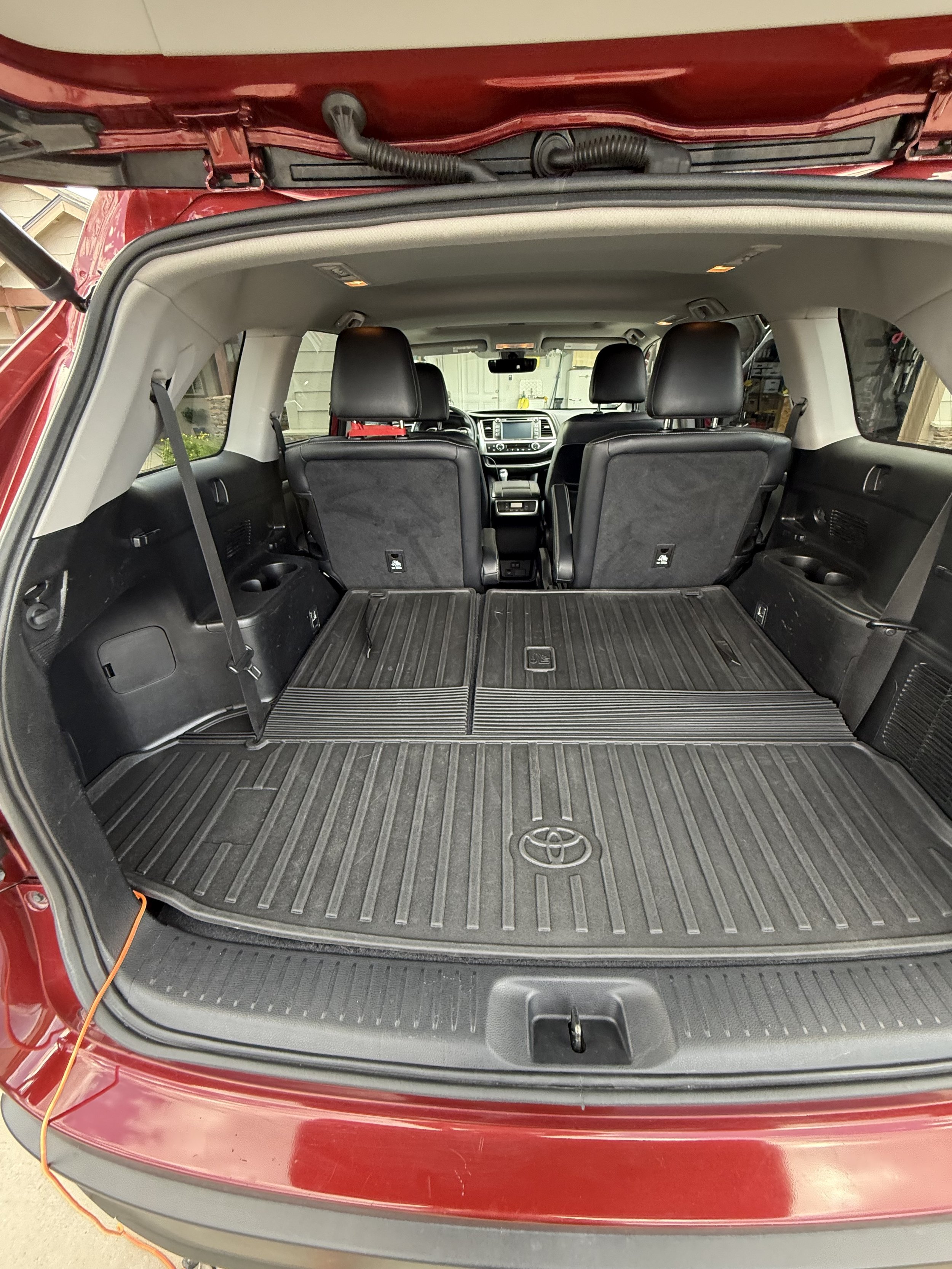 View of the open trunk of a red Toyota SUV showing the cargo area with black rubber mat and two rear seats, a dome light in the ceiling, and the interior dashboard visible through the rear window, with the vehicle parked in a garage.