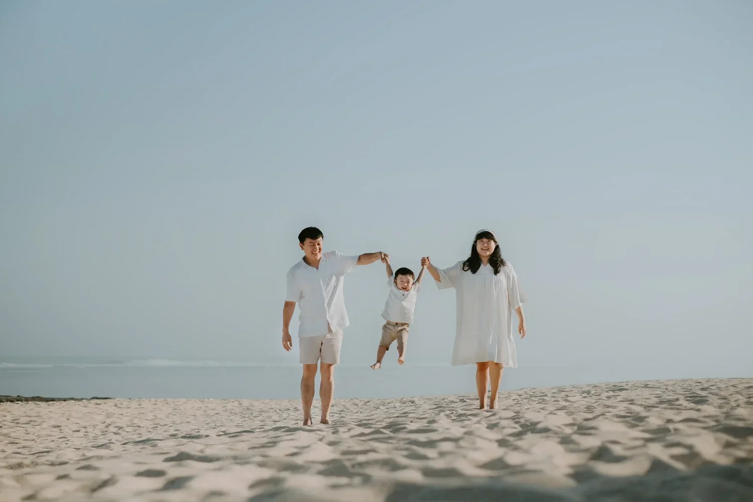 A family of three, parents and a child, playing on a sandy beach during daytime. The parents are holding the child's hands and lifting him in the air, all are smiling and wearing white clothing.