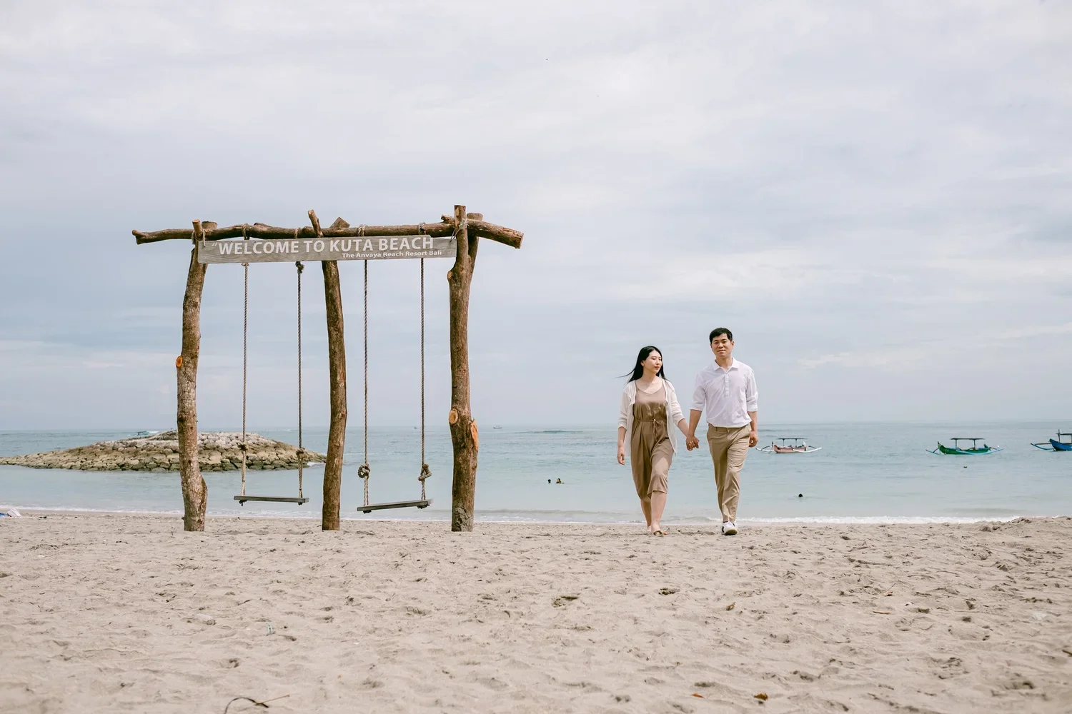 A couple holding hands walking on the beach near a wooden swing set with a sign that says "Welcome to Kuta Beach" under a cloudy sky, with boats in the water in the background.