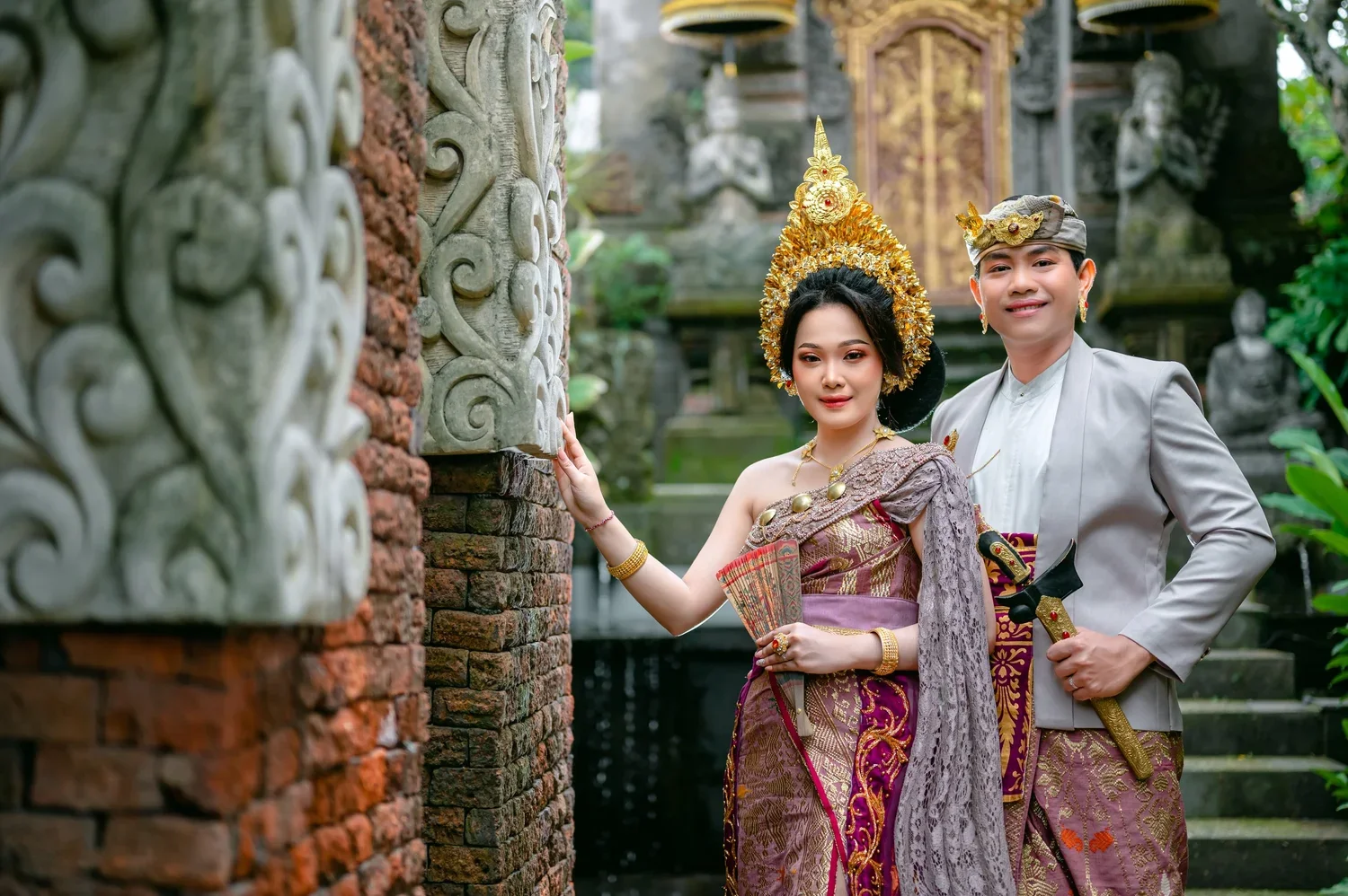 A woman and a man dressed in traditional Balinese attire, standing outdoors near an ornately carved stone structure with lush greenery in the background. The woman is wearing a purple and gold traditional outfit with gold jewelry and a headdress, hol