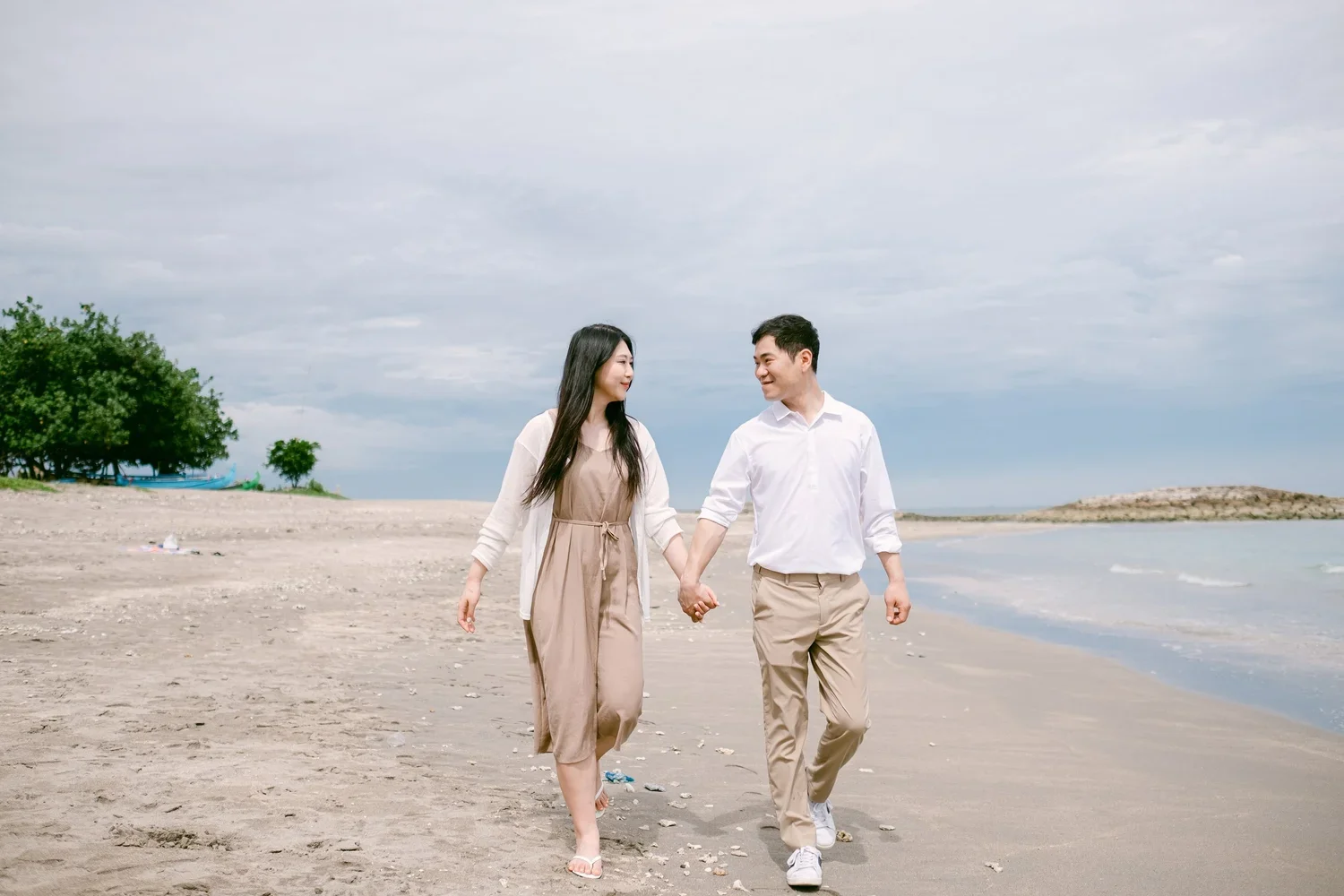 A young couple walking hand in hand on a beach, smiling at each other under a cloudy sky.