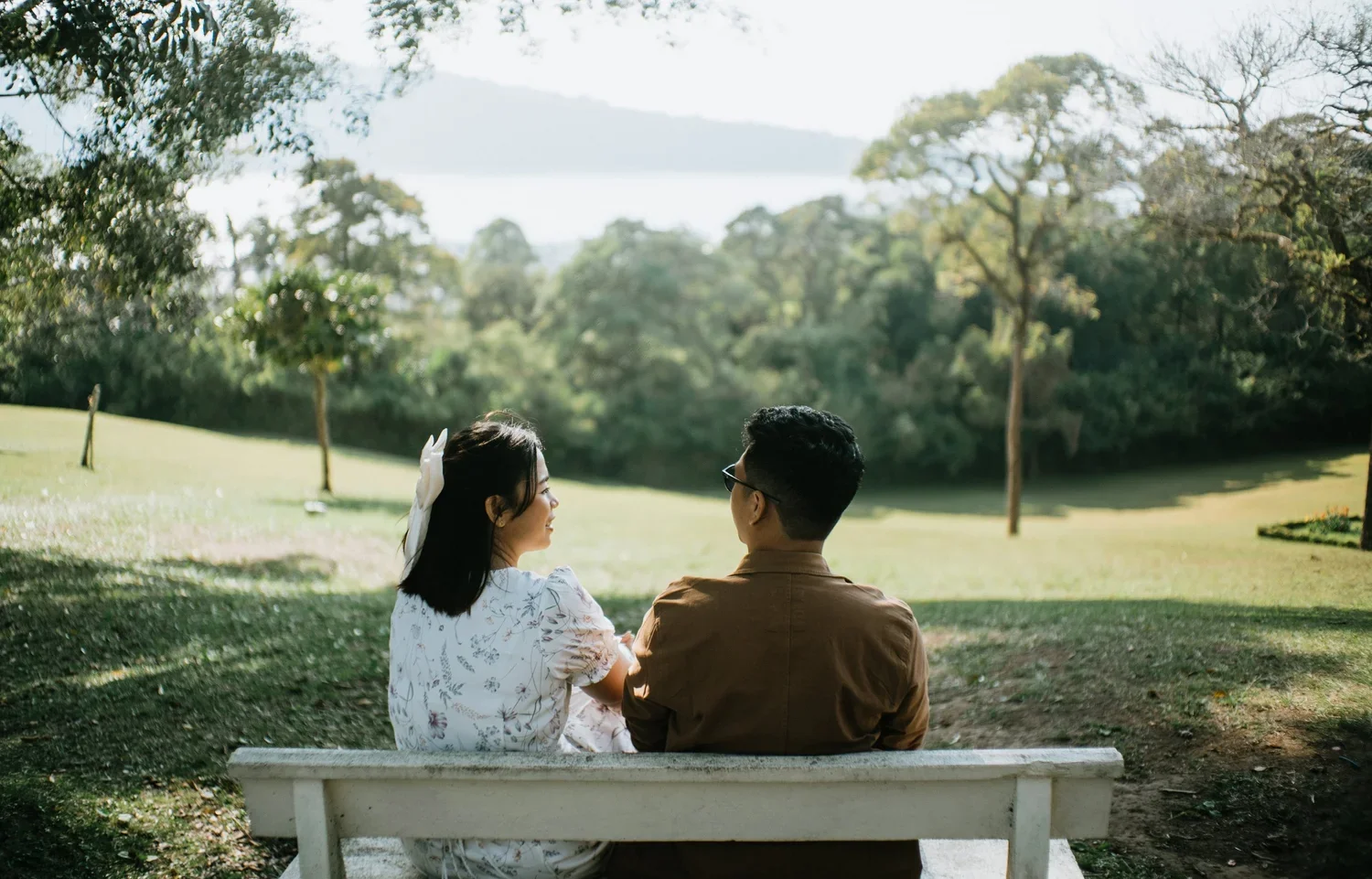 A man and woman sit on a white park bench outdoors, facing each other, in a lush green park with trees and a distant lake or river.
