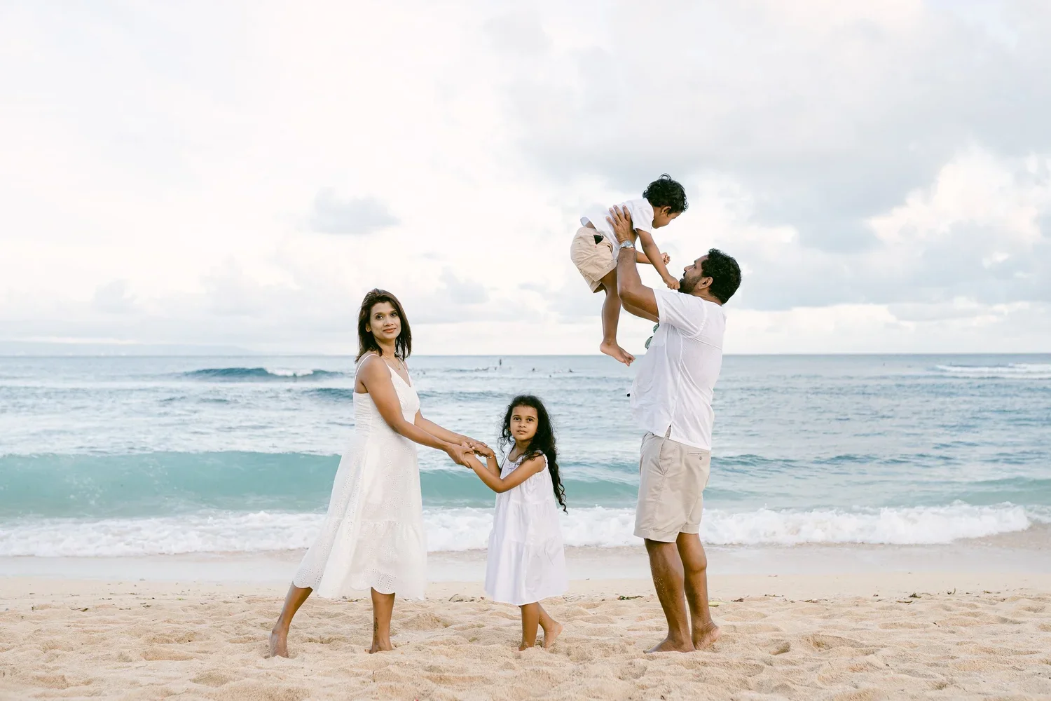 A family of four at the beach with the father lifting a young boy in the air, the mother holding the hand of a young girl, and all standing on the sand near the ocean with waves in the background.