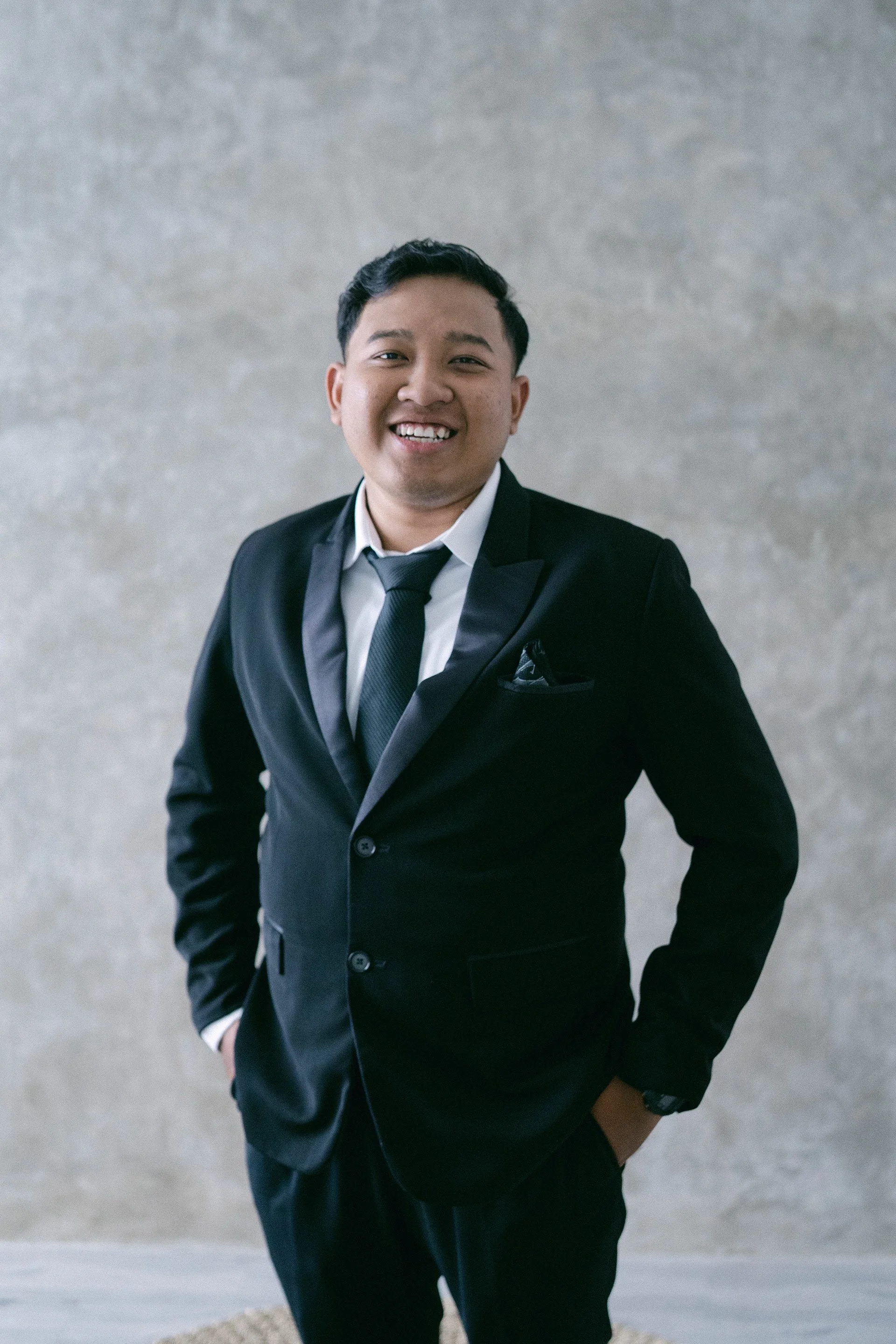 A man in a black suit and tie smiling at the camera, standing with one hand in his pocket against a plain background.