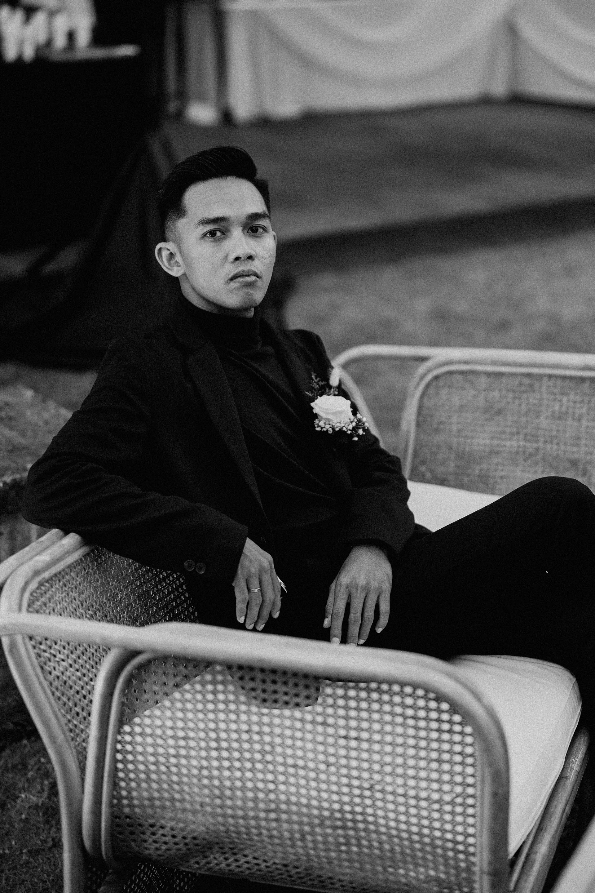 A young man with dark hair seated on a wicker chair, wearing a black suit and turtleneck, with a boutonniere on his lapel in a formal setting.